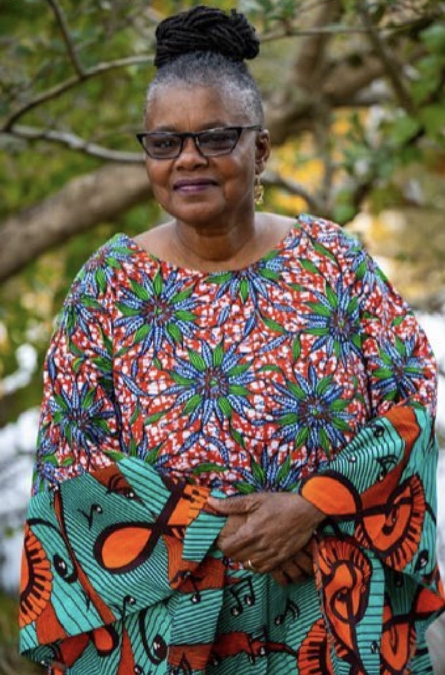 A woman with glasses and braided hair styled in an updo, wearing a colorful African-patterned dress with floral and musical note designs, standing outdoors with green foliage in the background.