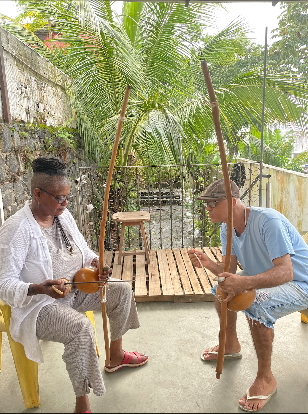 Two people sitting outdoors, playing traditional string instruments with long wooden necks and rounded gourds as resonators, under a large palm tree with a garden and stone wall in the background.