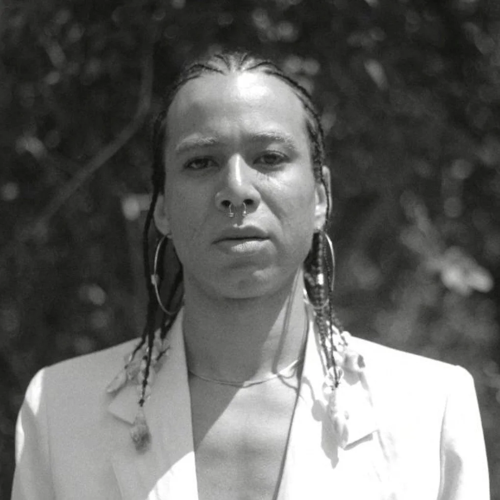 Black and white portrait of a woman with braided hair, hoop earrings, and a septum piercing, wearing a light-colored blazer, outdoors with blurred foliage in the background.