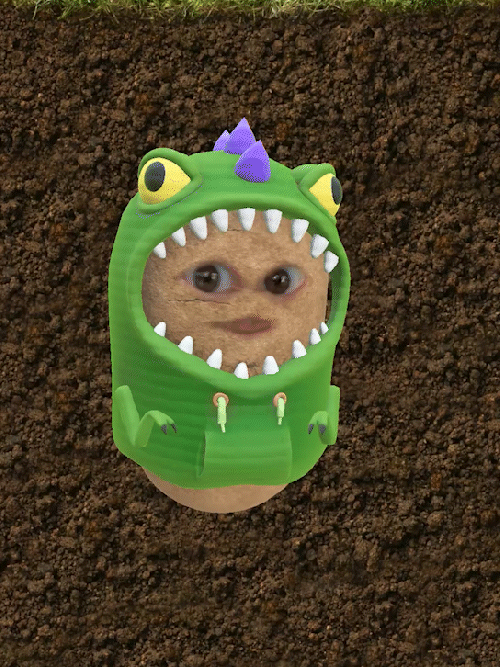 A potato with a dinosaur costume featuring cartoonish eyes, purple horns, and sharp teeth, with a person's face inside, set against a dirt background.
