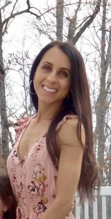 A woman with long dark hair smiling outdoors among leafless trees, wearing a pink floral dress.