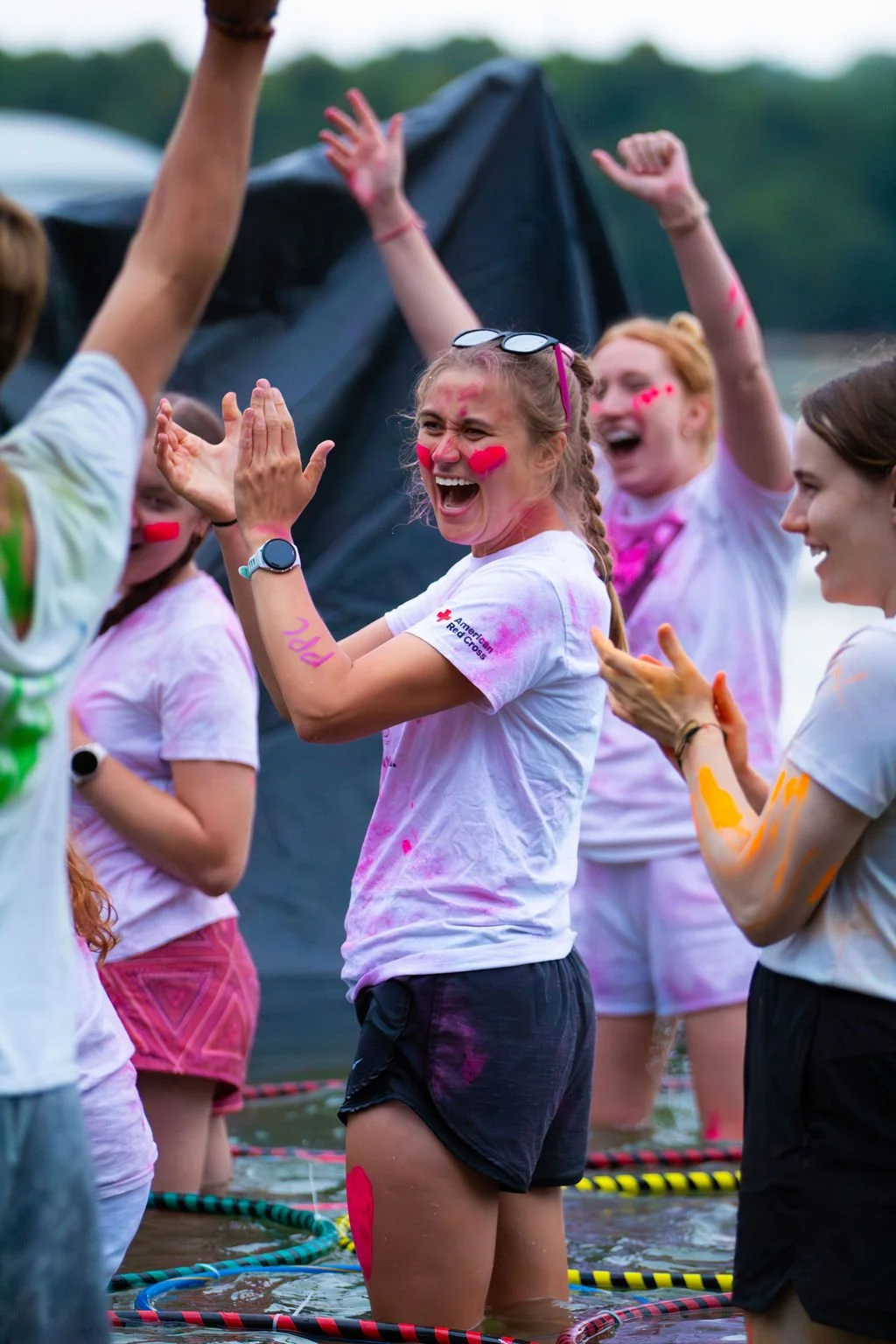 Group of women celebrating, covered in colorful powder at an outdoor event, some wearing white t-shirts with a Red Cross logo, standing in water with floating ropes.