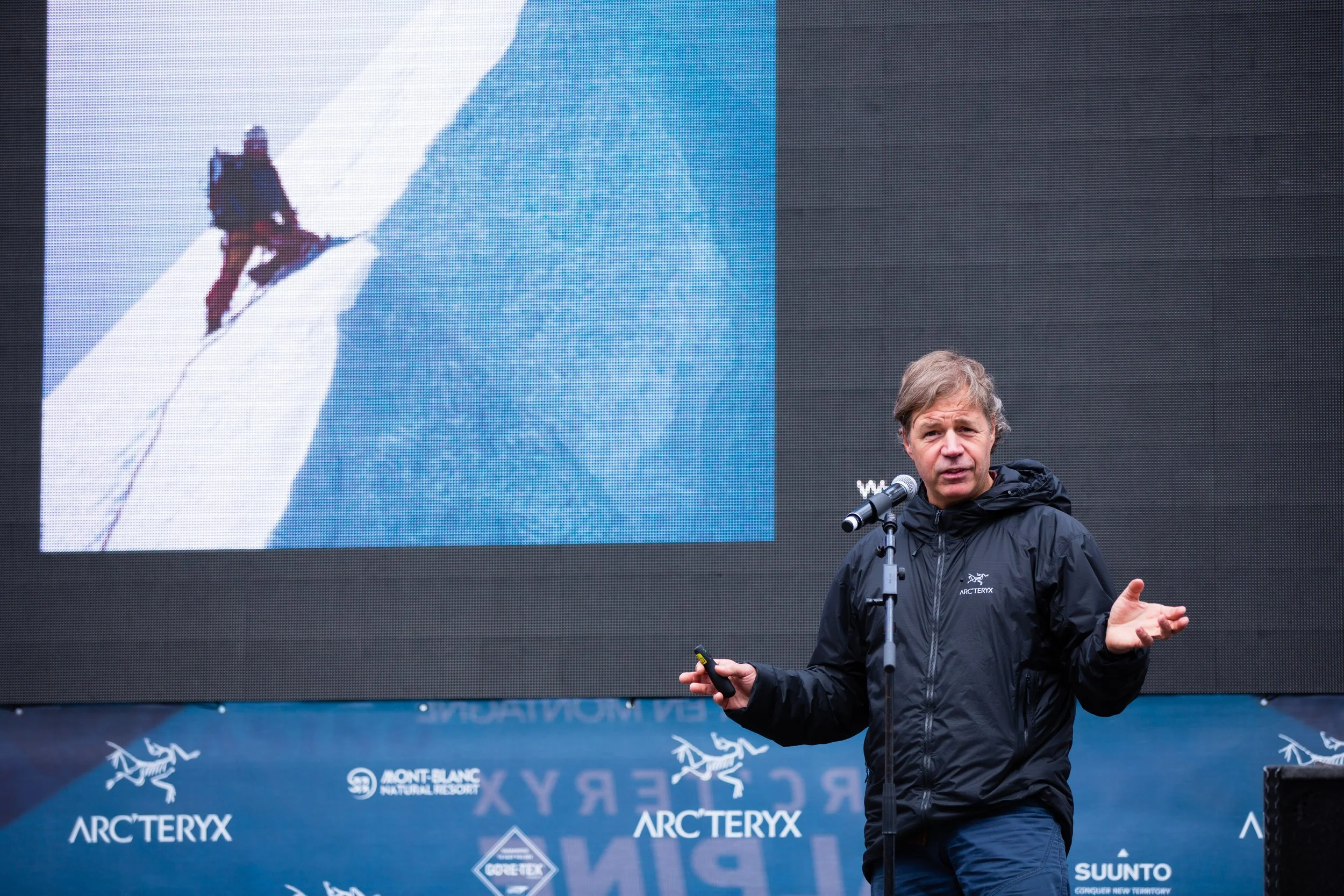 A man in a black Arc'teryx jacket speaking at a podium with a microphone, during a presentation or conference. Behind him is a large digital screen displaying an image of a person climbing a snowy mountain, with logos of Mont Blanc, Arc'teryx, and other sponsors.