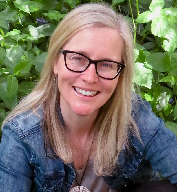 A smiling woman with blonde hair and glasses, wearing a denim jacket, posing outdoors in front of green foliage.