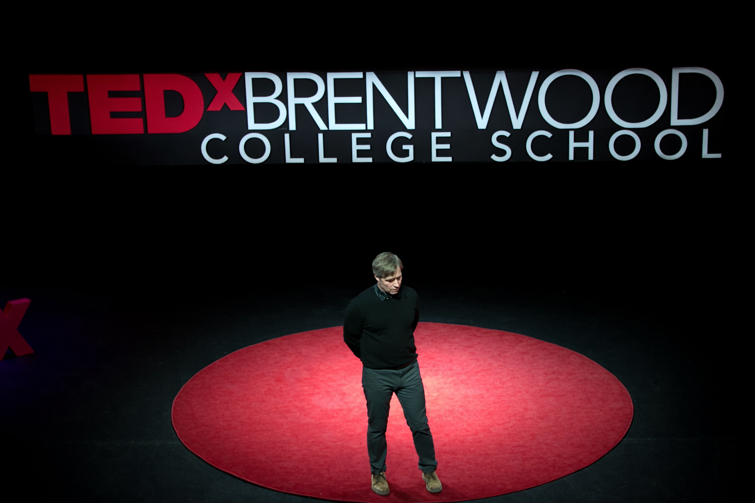 A man stands on a red circular carpet on a stage with a large sign that reads 'TEDxBrentwood College School' in the background.