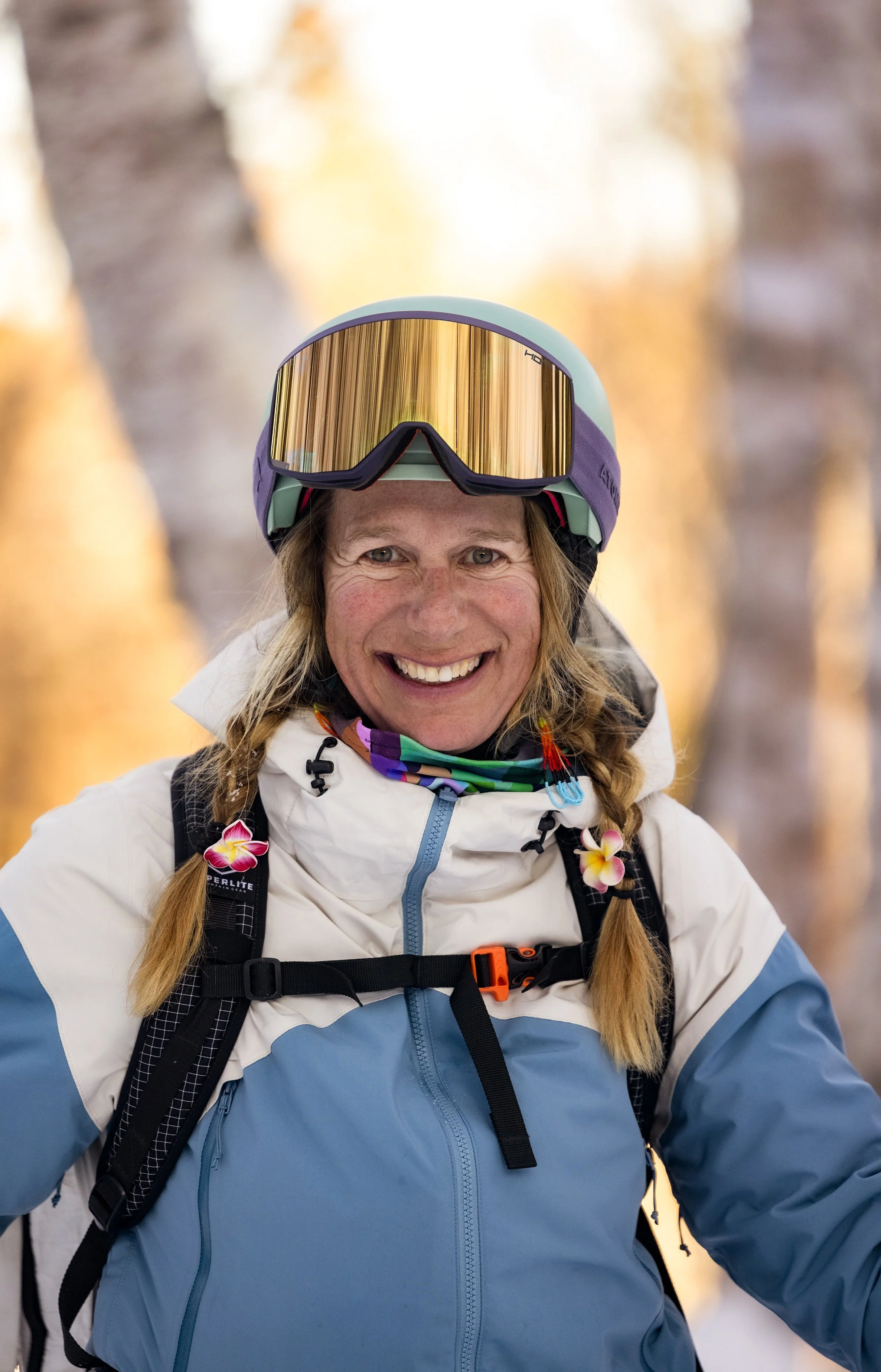 A woman smiling outdoors in winter gear, wearing ski goggles and a jacket, with two braided pigtails decorated with pink and yellow flowers, in a forested area with blurred trees in the background.