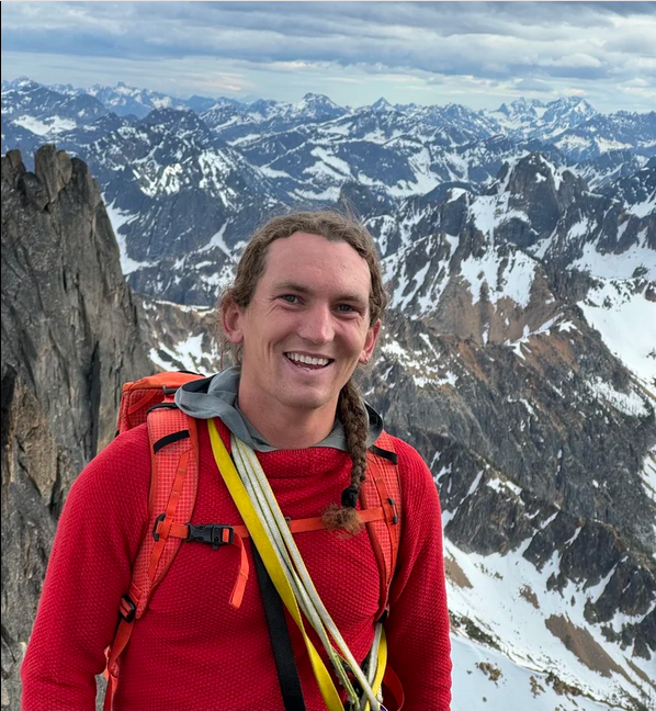 Smiling man in red outdoor gear and backpack hiking in snowy mountain terrain with rugged peaks in the background.