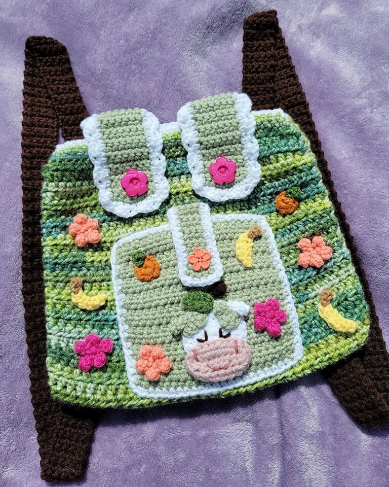 Crocheted backpack with cow design and floral decorations on a purple background.