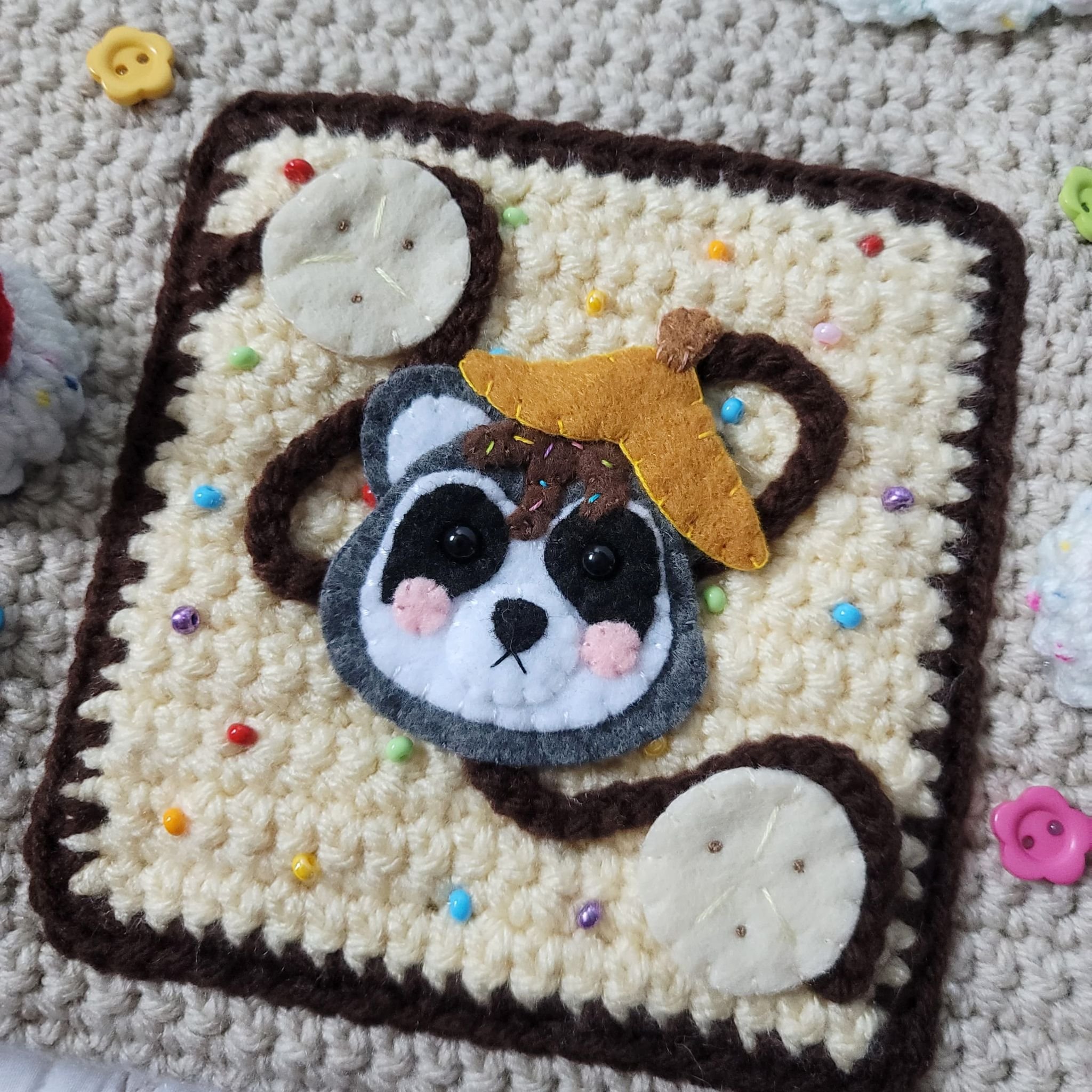 A crocheted square with a felt raccoon face adorned with a yellow felt hat. The background is sprinkled with colorful beads, resembling sprinkles, and includes circular felt pieces that look like scoops of vanilla ice cream. A pink button in the shap