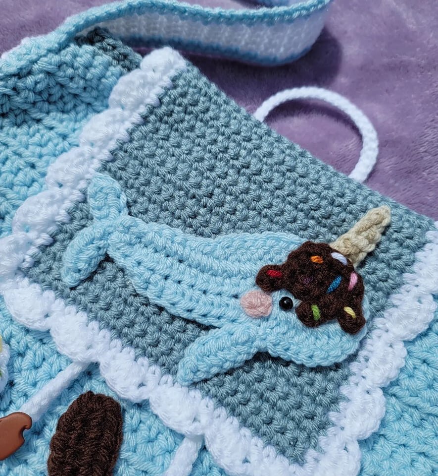 Crocheted narwhal with ice cream cone hat, on a blue and gray textured background, with scalloped edges.
