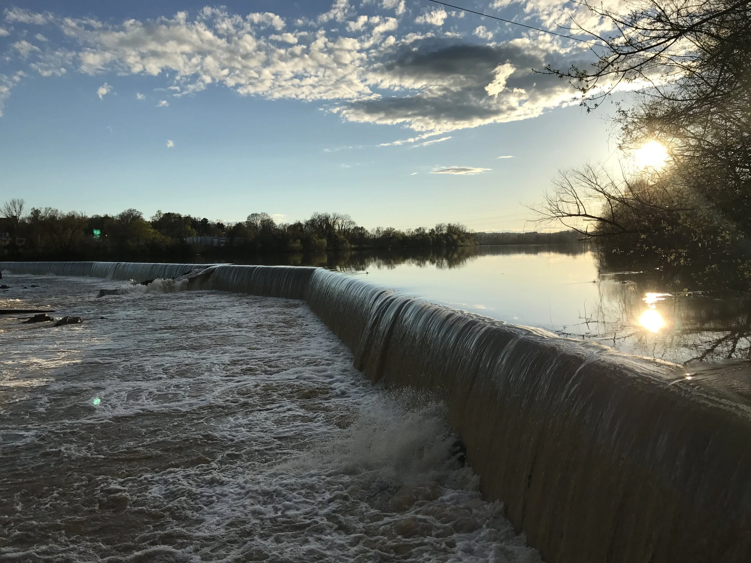 A river with a waterfall at sunset, reflecting the sky and trees along the bank.