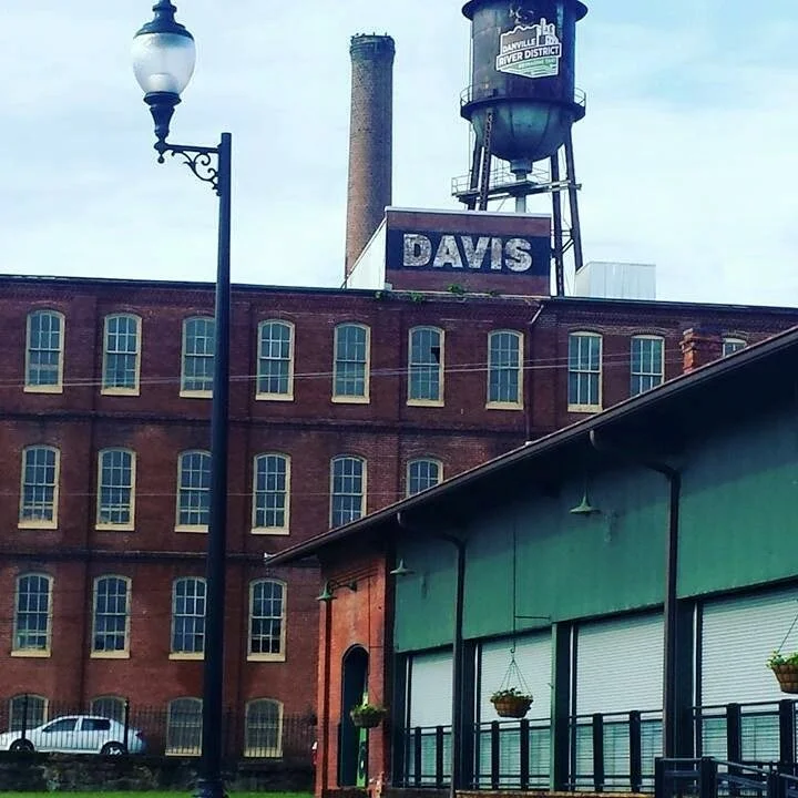 A historic brick building with multiple windows, a green building attached, and a water tower with a sign that says 'Davis' and a smaller sign indicating 'Davisville River District' in the background.