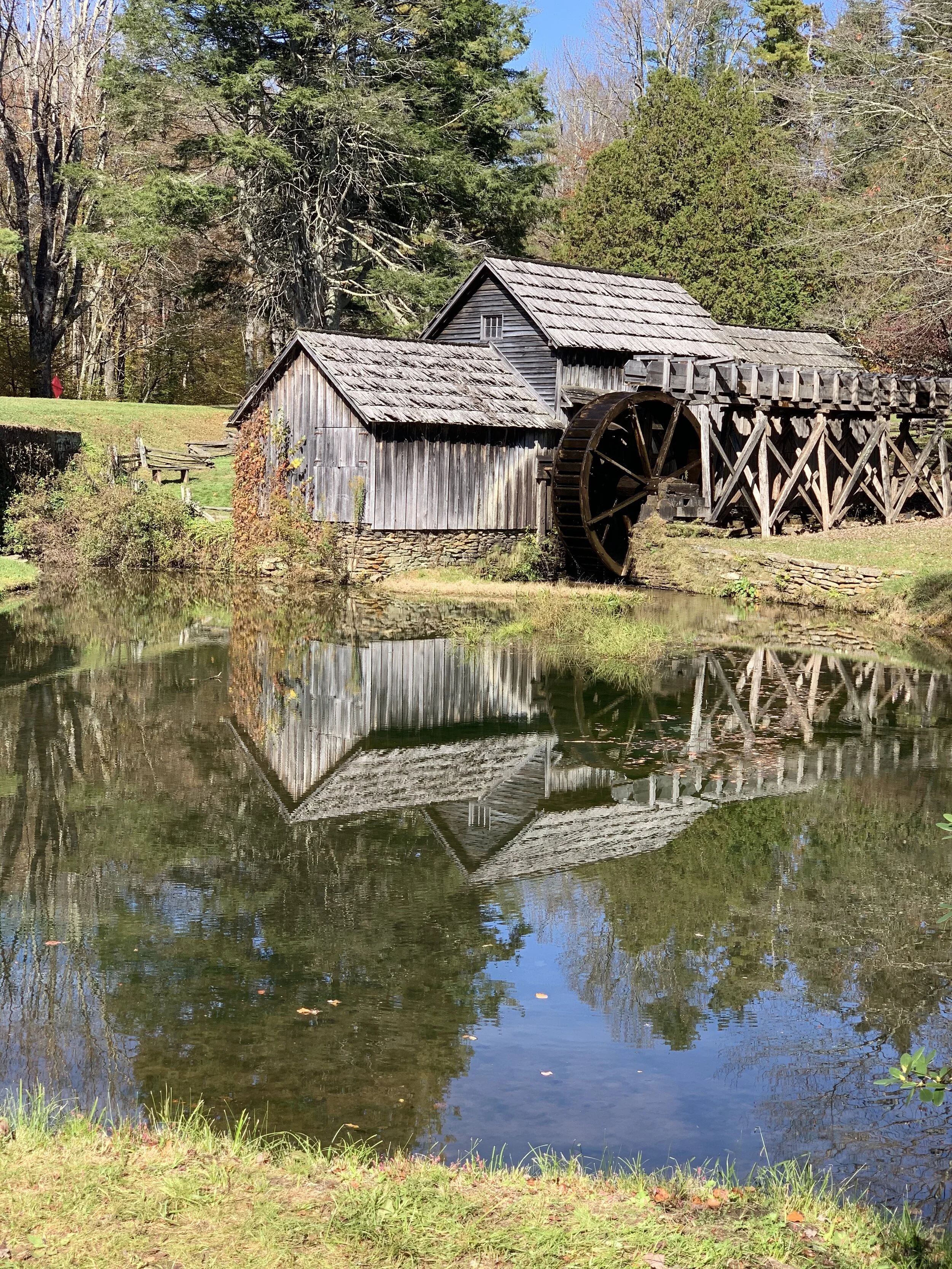 A historic wooden watermill beside a pond, with its reflection visible in the water, surrounded by trees and grass.