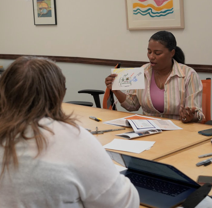 Two individuals are seated at a table in a meeting room. One person is holding and explaining a diagram, while the other listens. Laptops, papers, and stationery are spread on the table.