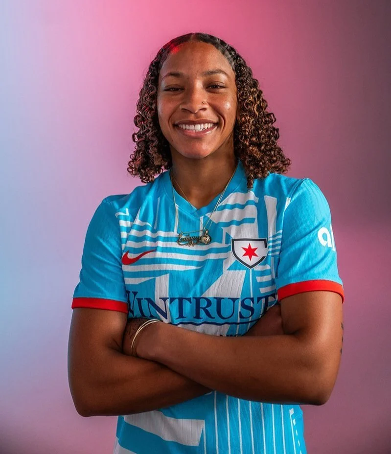 Soccer player Leilanne Nesbeth with curly hair wearing a blue sports jersey with red accents and a white and blue emblem, standing with arms crossed against a pink and purple gradient background.