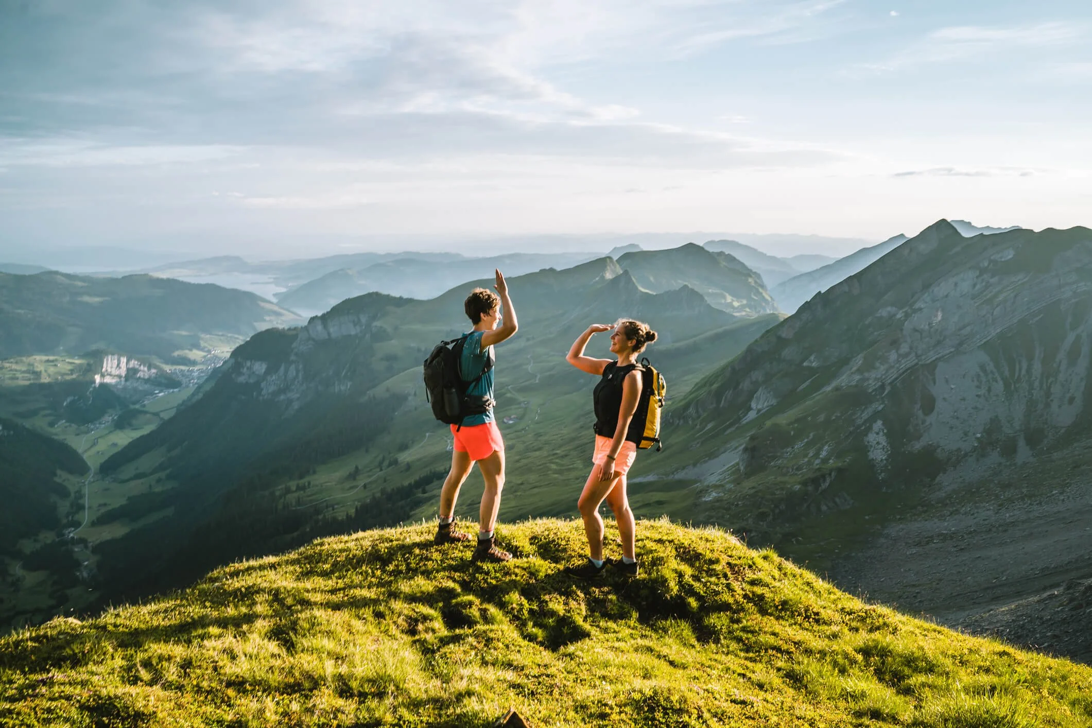 Deux randonneuses saluant au sommet d'une colline avec vue sur des montagnes verdoyantes et un ciel partiellement nuageux.