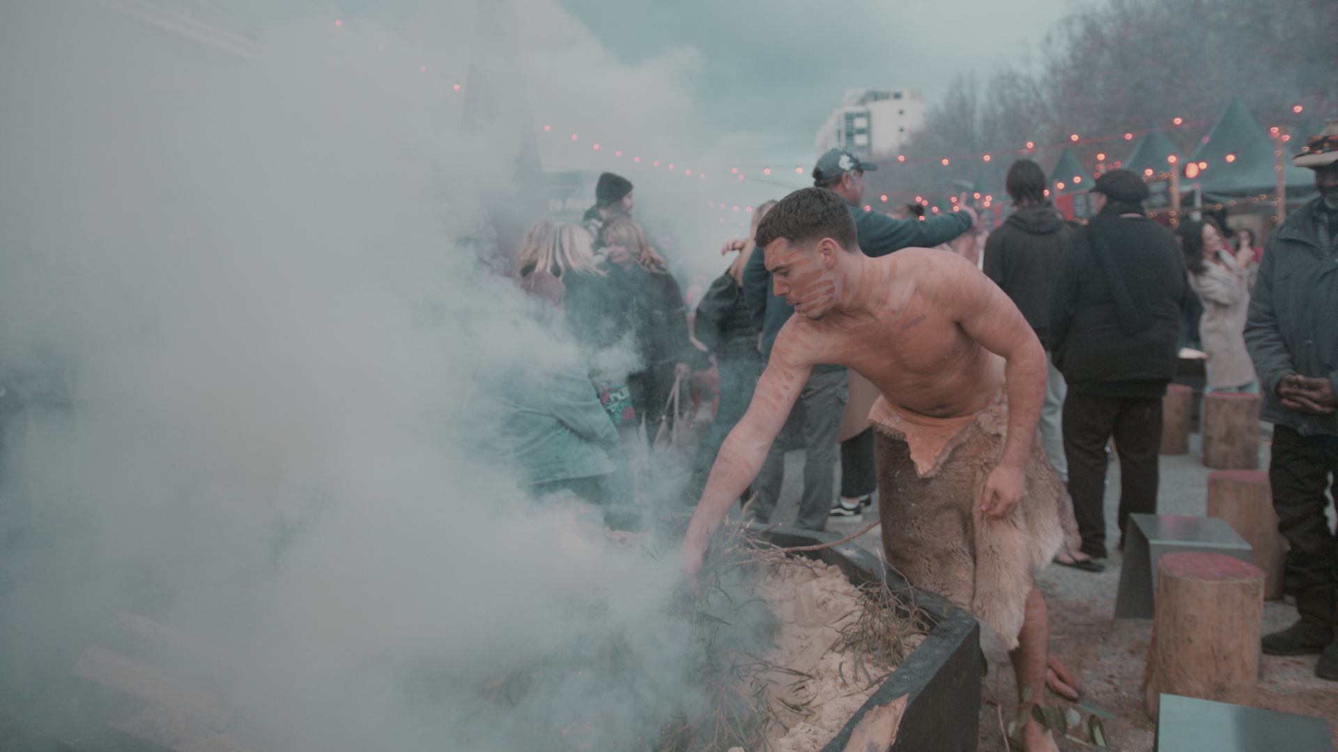 A performer tending smoking branches as visitors gather nearby at The Gathering, Winter Feast.