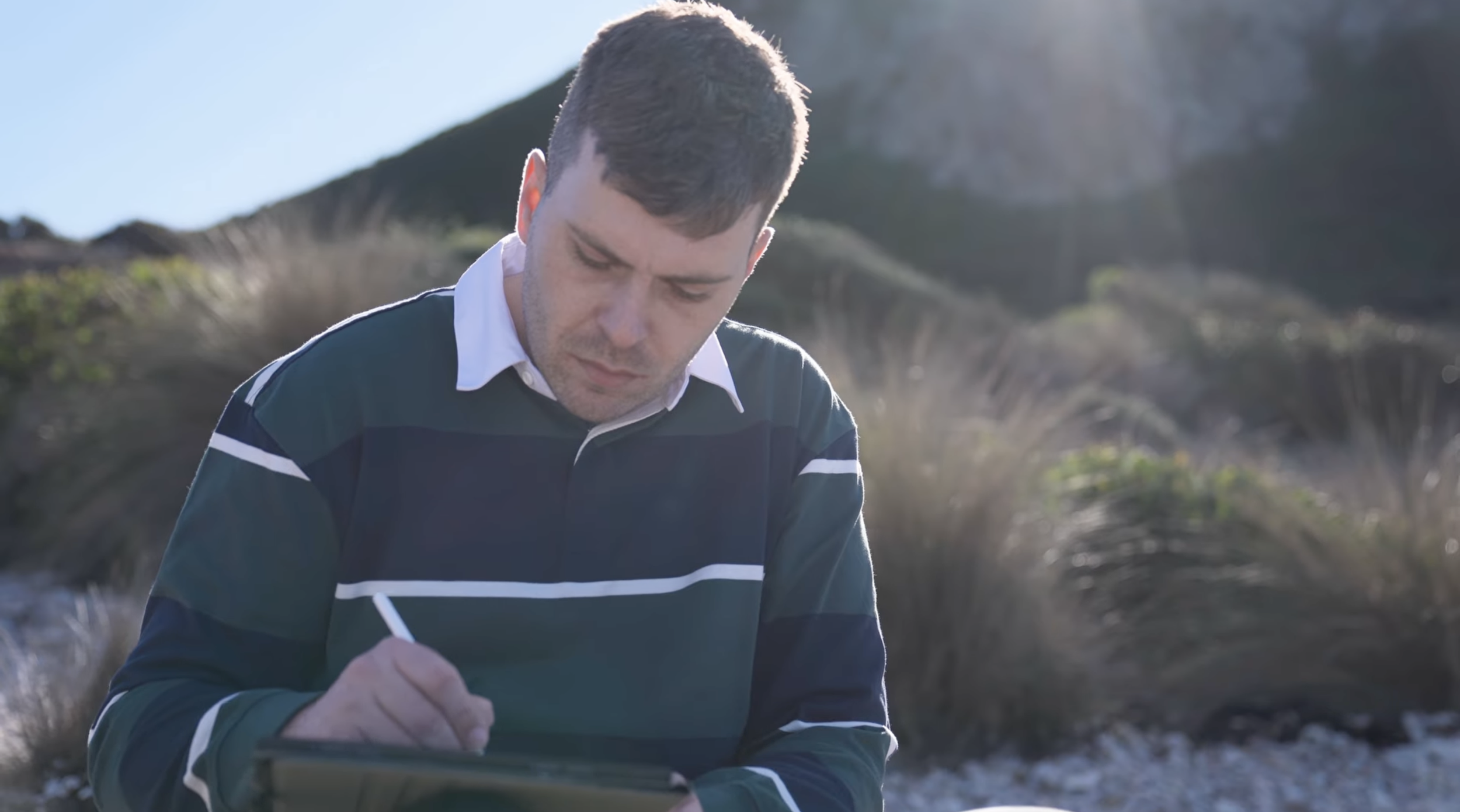 Palawa artist Caleb Nichols-Mansell drawing on an iPad while seated on Country among grasses and shrubs.
