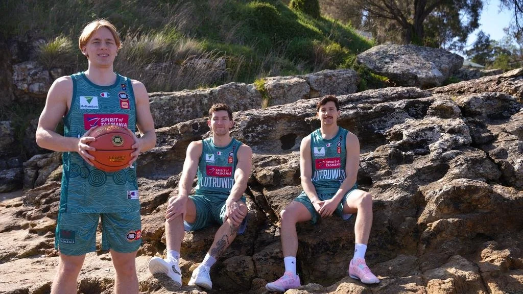Three Tasmania JackJumpers players wearing teal Indigenous Round uniforms, one standing with a basketball and two seated on rocks.