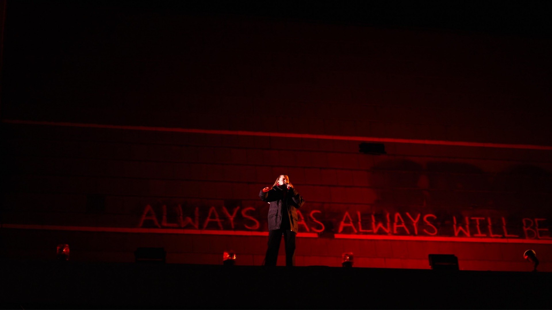 DENNI singing on a dark roof awning illuminated by red lighting at Dark Mofo's 2026 Night Mass. The background features the phrase "ALWAYS WAS ALWAYS WILL BE" as she performs 'Out Here'.
