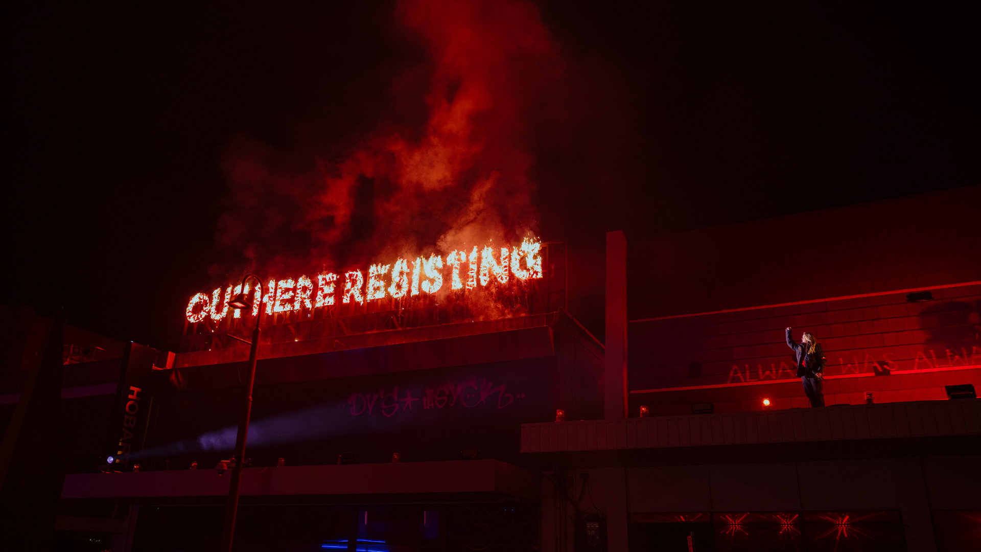 A building with a fiery sign reading 'OUT HERE RESISTING' on the roof, with flames and smoke rising from the sign at night, and DENNI standing on the roof awning after her Night Mass performance at Dark Mofo 2026.