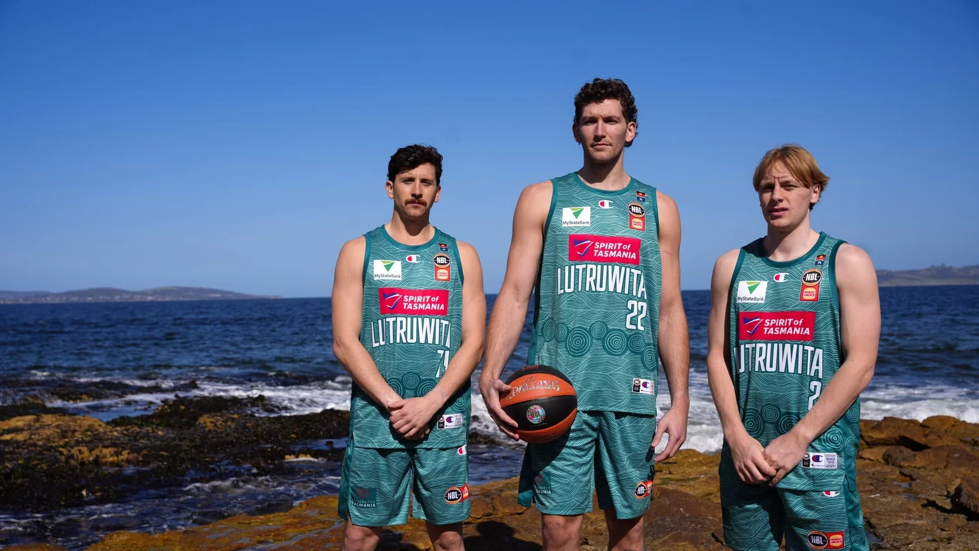 Three Tasmania JackJumpers players wearing teal Indigenous Round jerseys and shorts on a rocky shoreline.
