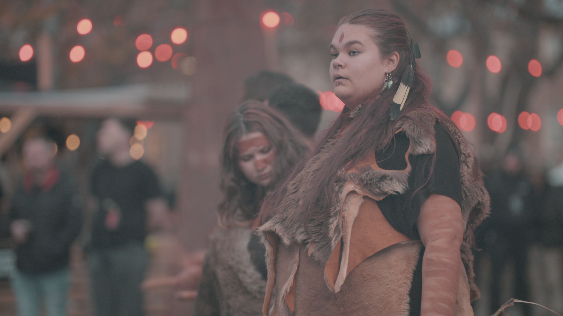 Two Pakana Kanaplila performers wearing skin garments during The Gathering, with red festoon lights behind them.