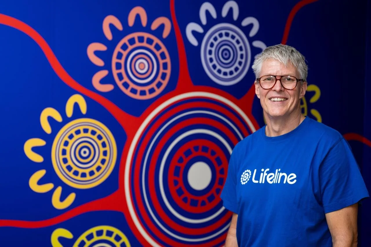 Graham Strong in a blue Lifeline shirt standing in front of the feature wall with orange journey lines and concentric motifs.