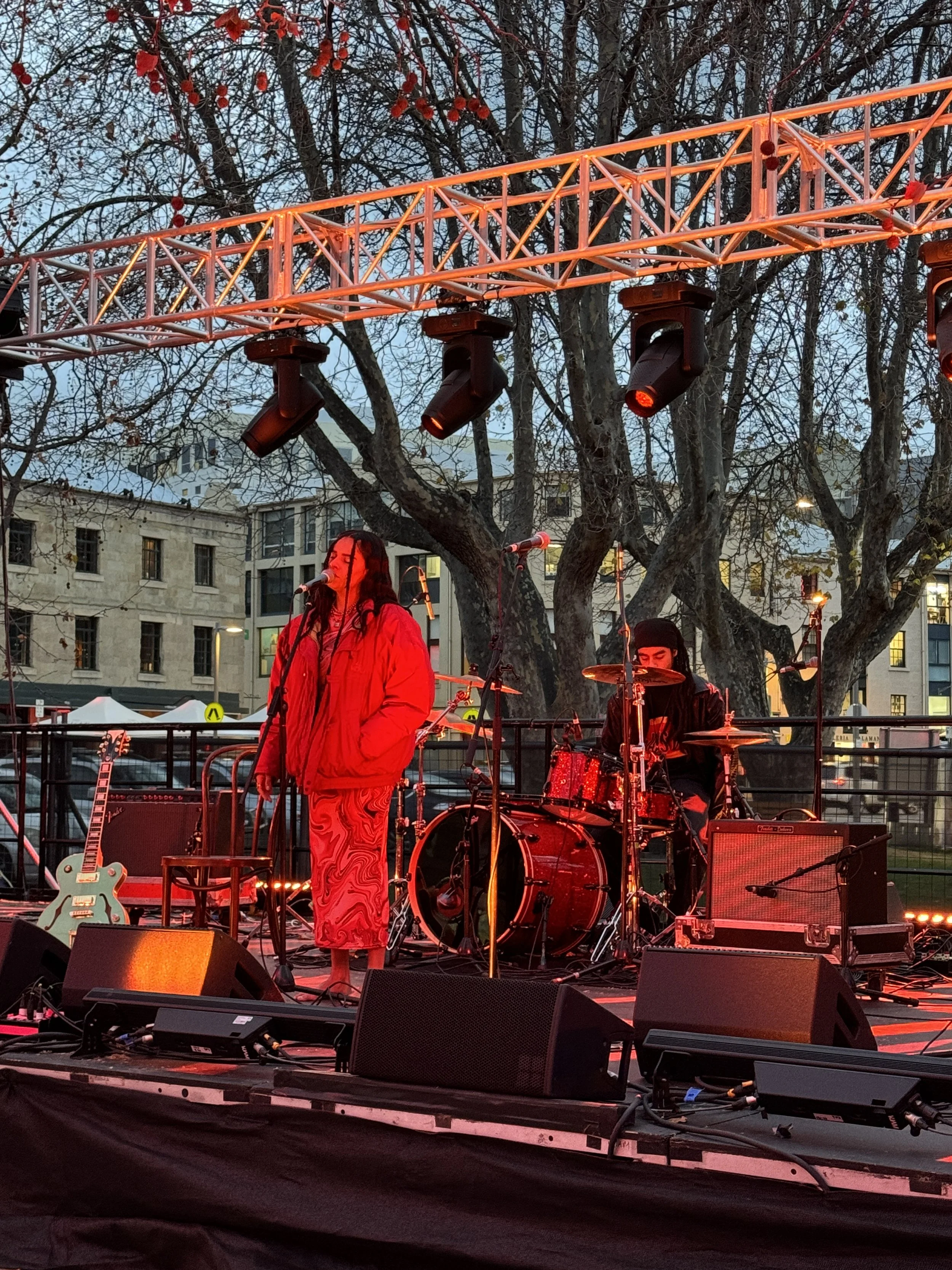 LEO stands singing with their eyes shut under red lighting on the main stage at The Gathering, Winter Feast 2025. LEO is at the front of the stage with their partner behind them on the drums.