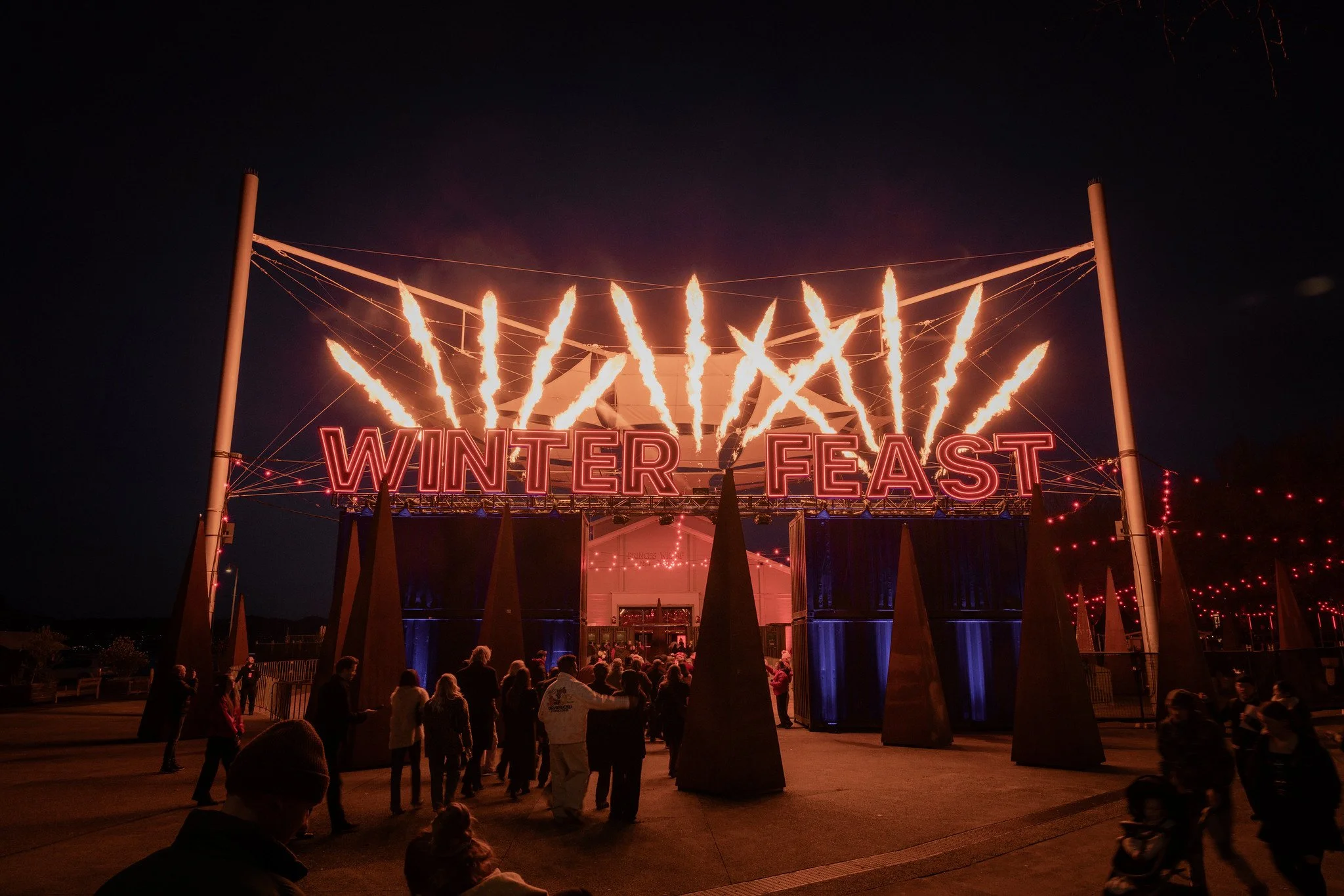 Visitors entering Winter Feast beneath a red illuminated sign and jets of flame at Dark Mofo in Hobart.