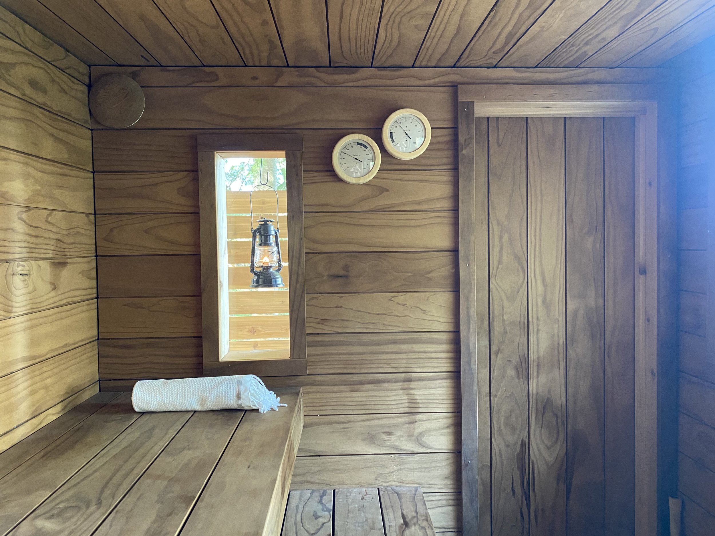 Interior of a wooden sauna with a bench, a rolled-up towel, a small window, and two wall gauges.
