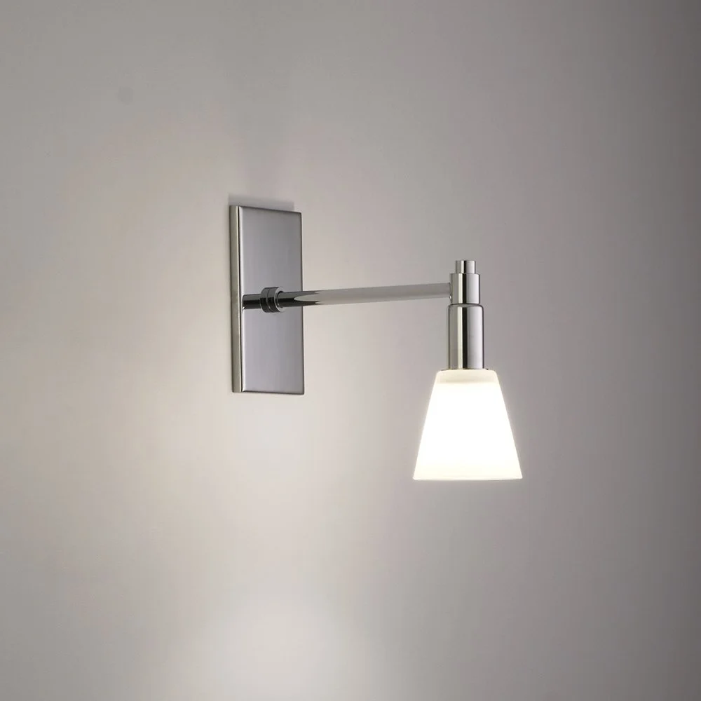 an illuminated sconce with polished chrome vertical mounting plate and a long central stem holding a downward white conical glass luminary
