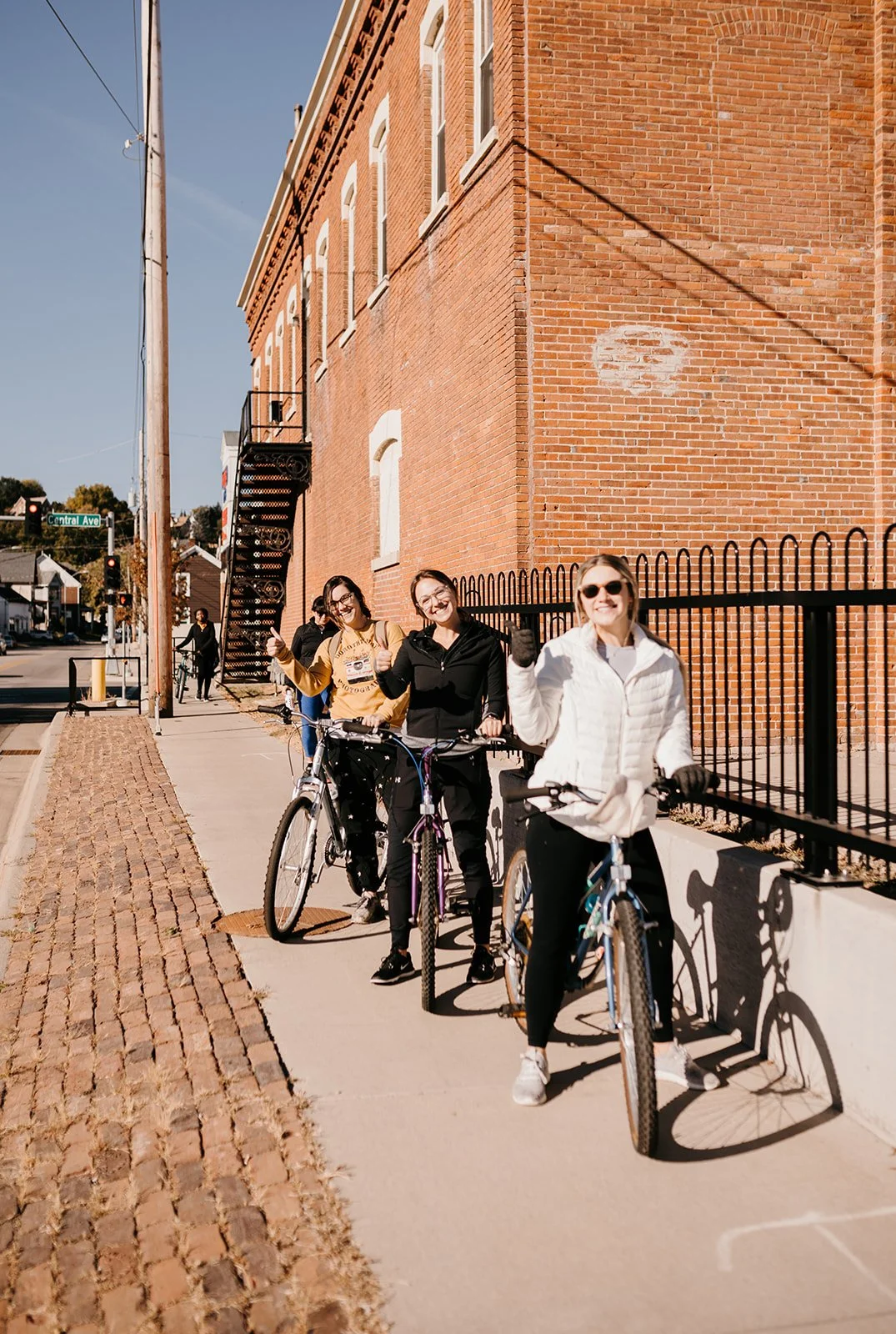 Four people with bicycles standing on sidewalk beside a tall brick building, all smiling and giving thumbs up.