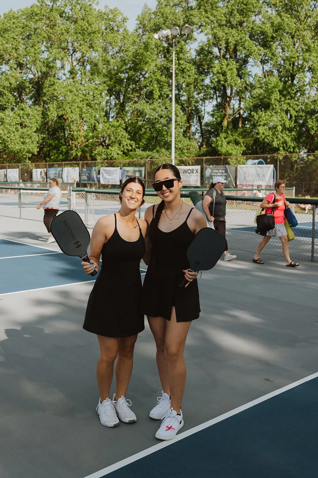 Two women in black athletic dresses holding tennis paddles on a tennis court, smiling for the camera. Several people walk past in the background on a sunny day with lush green trees.