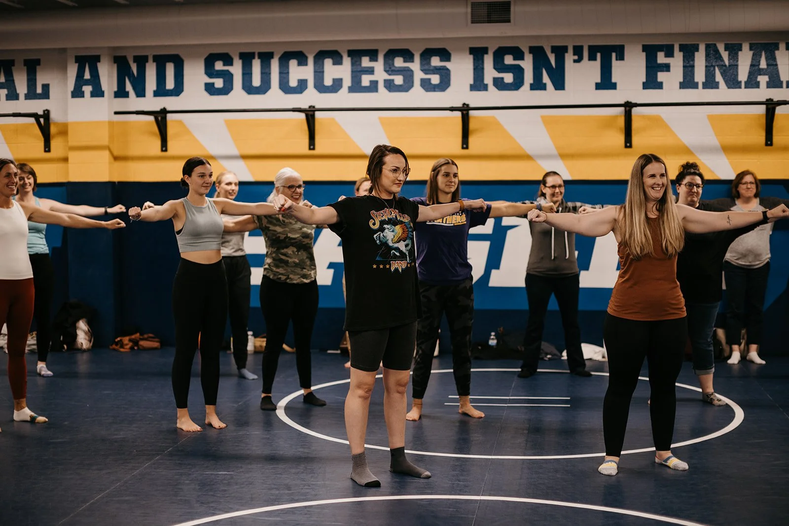 Group of women practicing yoga or stretching in a gym, with arms extended sideways, some smiling, on a blue floor with a yellow and blue wall in the background with motivational text.
