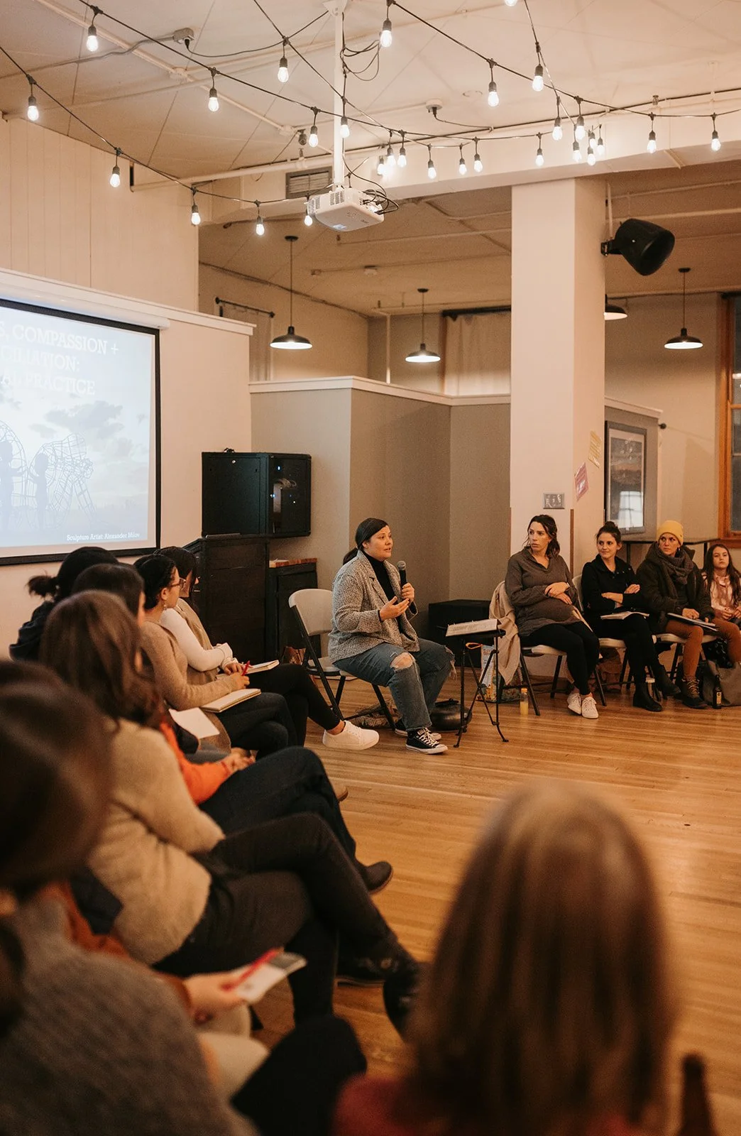 A woman in a gray blazer holding a microphone and speaking to an audience in a room decorated with string lights, with several women seated and listening attentively.