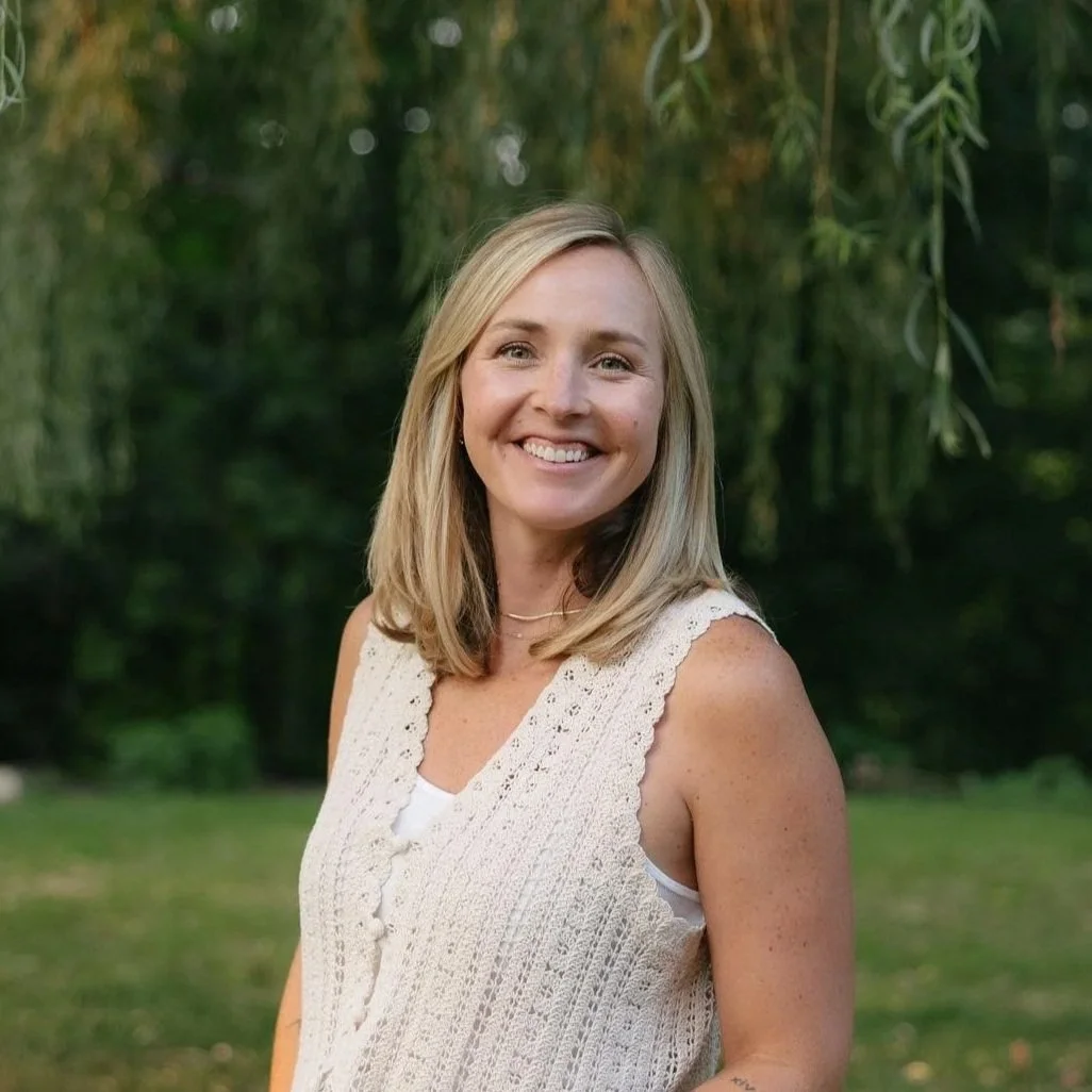 A smiling woman with blonde hair standing outdoors in front of greenery.