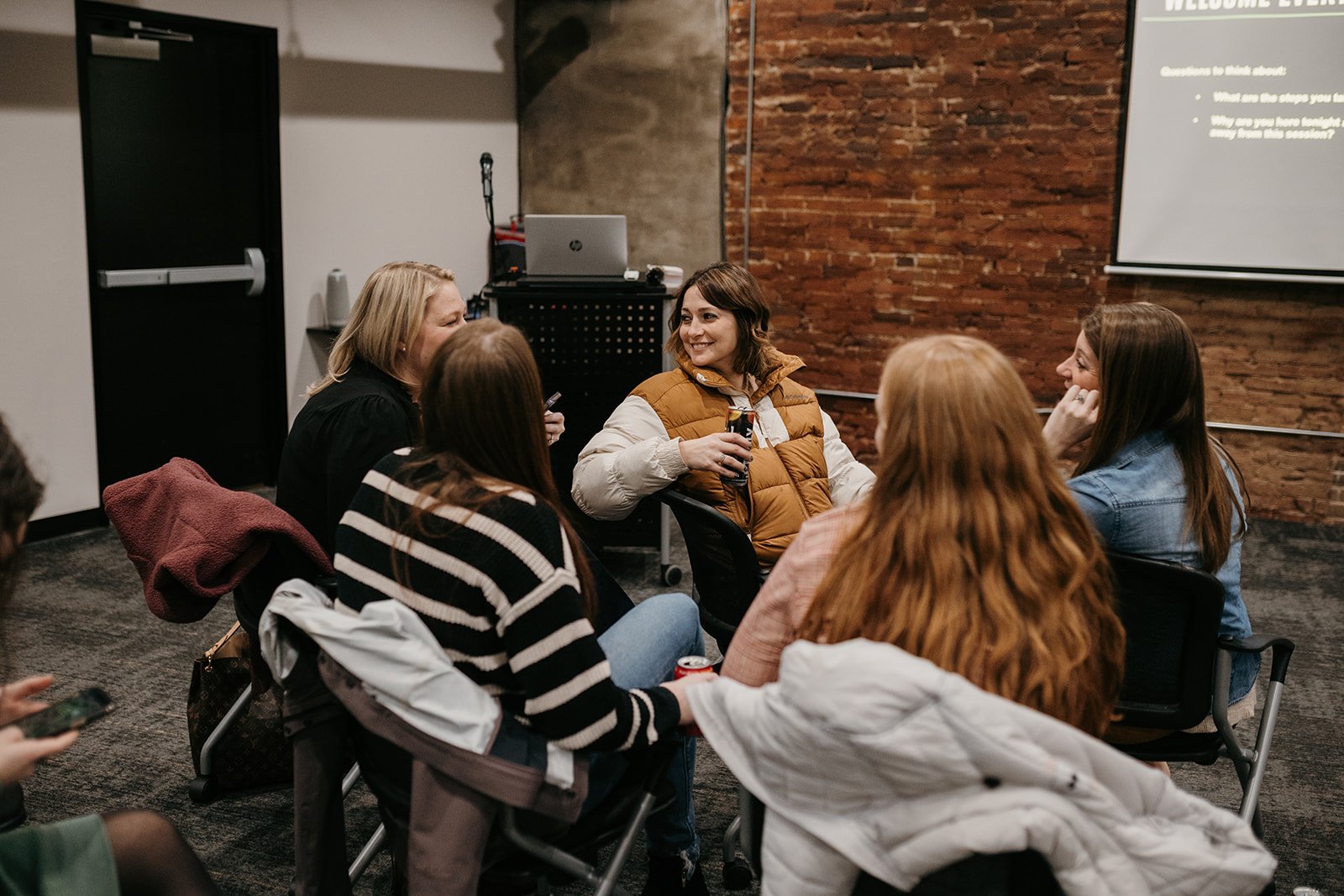 A group of women sitting in a circle during a discussion in a room with a brick wall, some holding drinks and smiling.