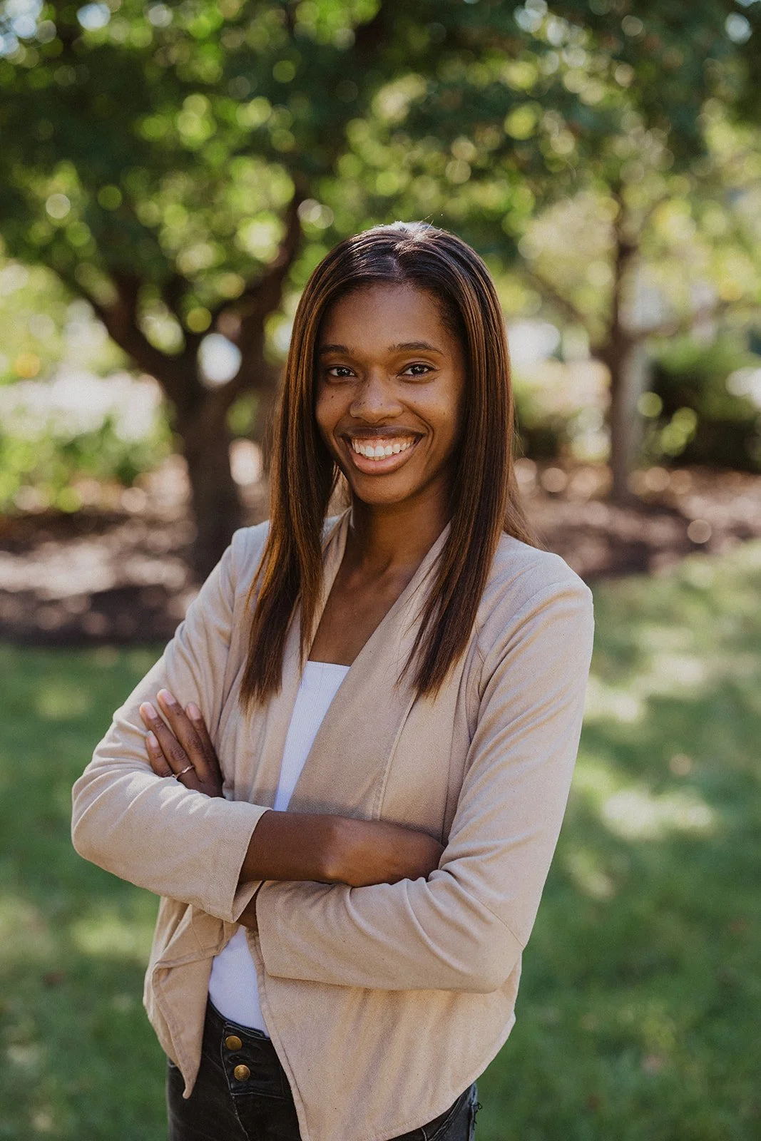 A smiling young woman with long straight hair, standing outdoors in front of a tree with green leaves. She is wearing a beige blazer over a white top and has her arms crossed.