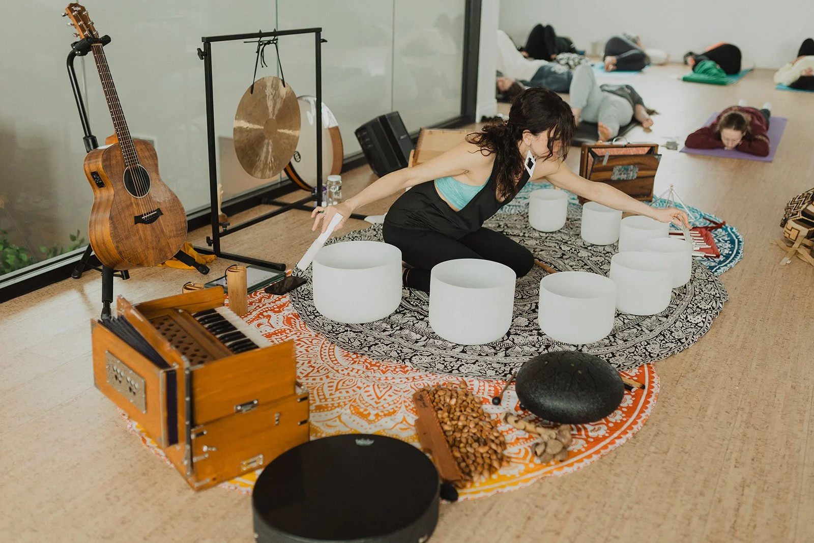 A group of people practicing yoga and playing musical instruments in a spacious room with large windows. A woman is sitting on a patterned rug, reaching toward a white singing bowl, surrounded by other bowls, a gong, and various percussion instruments. In the background, others are lying on yoga mats, resting or meditating.
