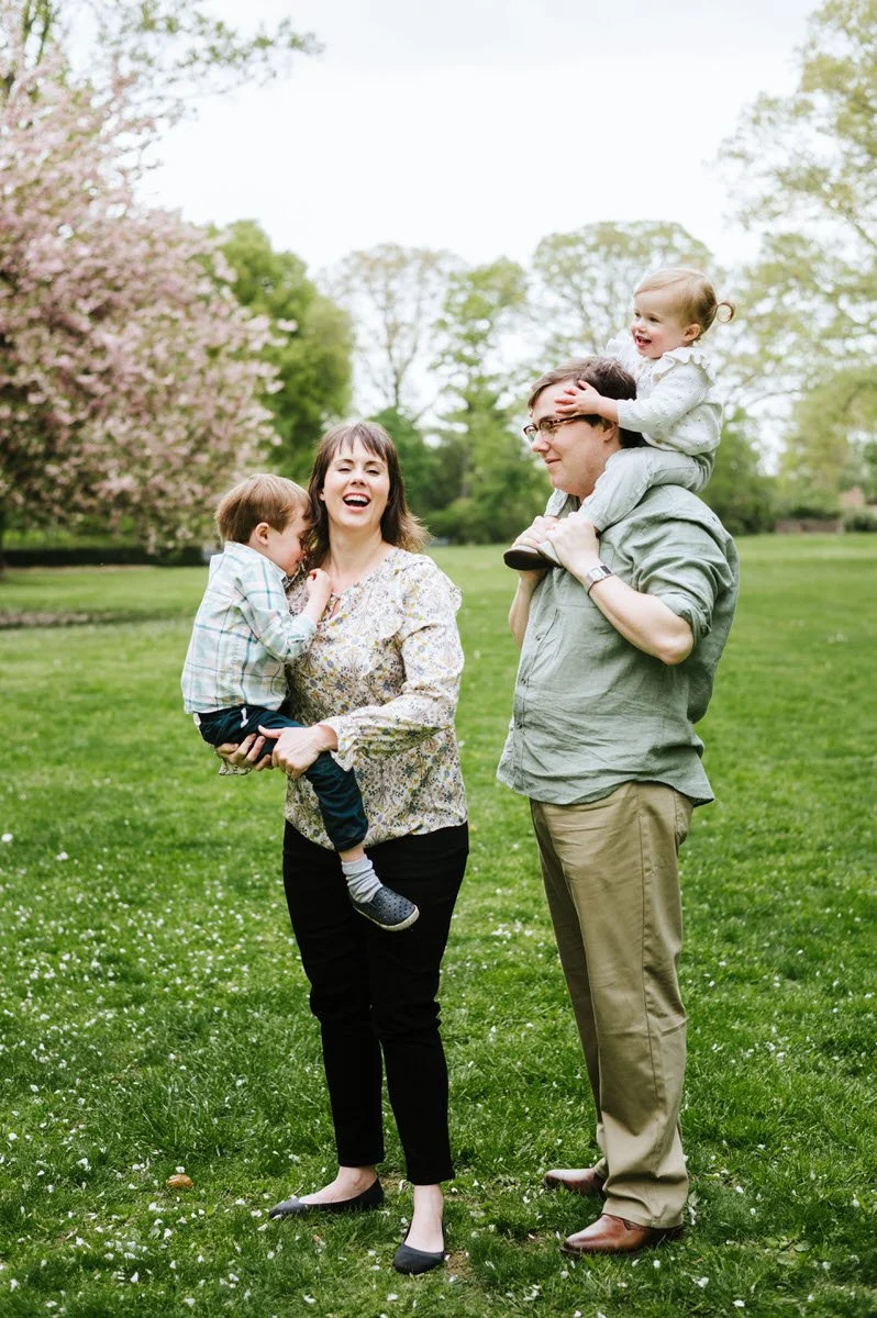 A family enjoying a day outdoors in a park with green grass and flowering trees. The woman is holding a young boy, and a man is carrying a young girl on his shoulders. They are all smiling and appear to be happy.