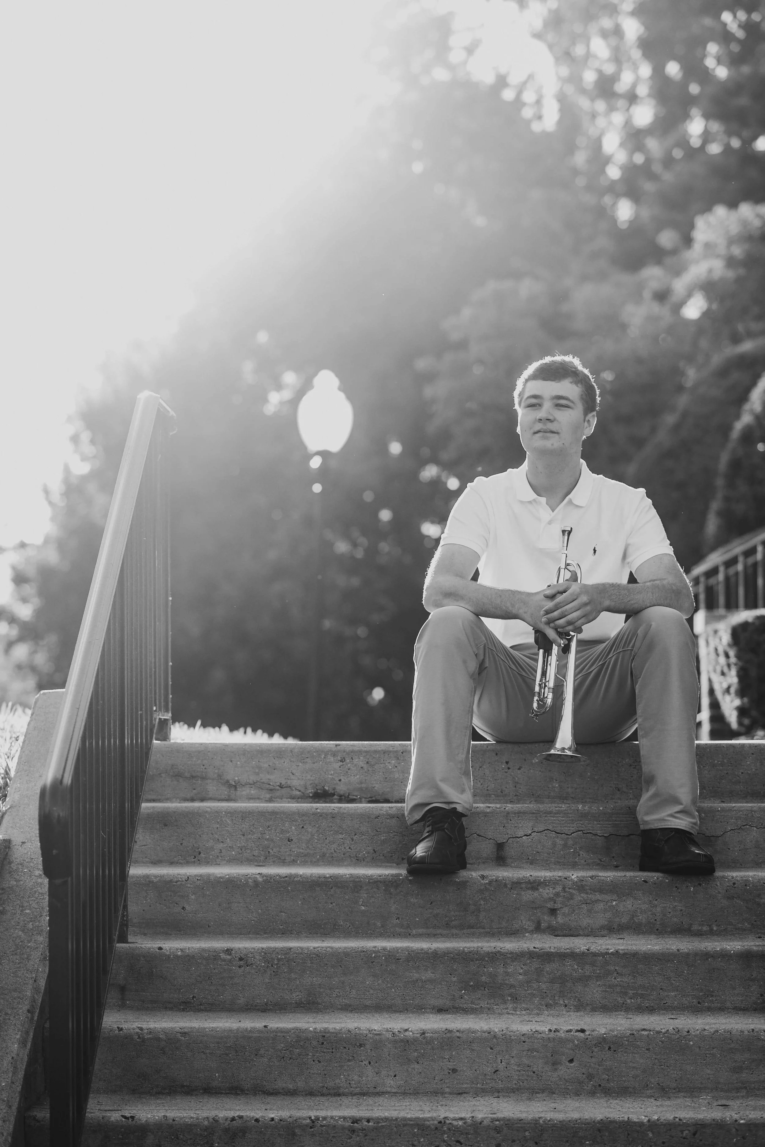 Young man sitting on concrete steps holding a trumpet, outdoors with trees and a lamppost in the background, backlit by the sun, in black and white.