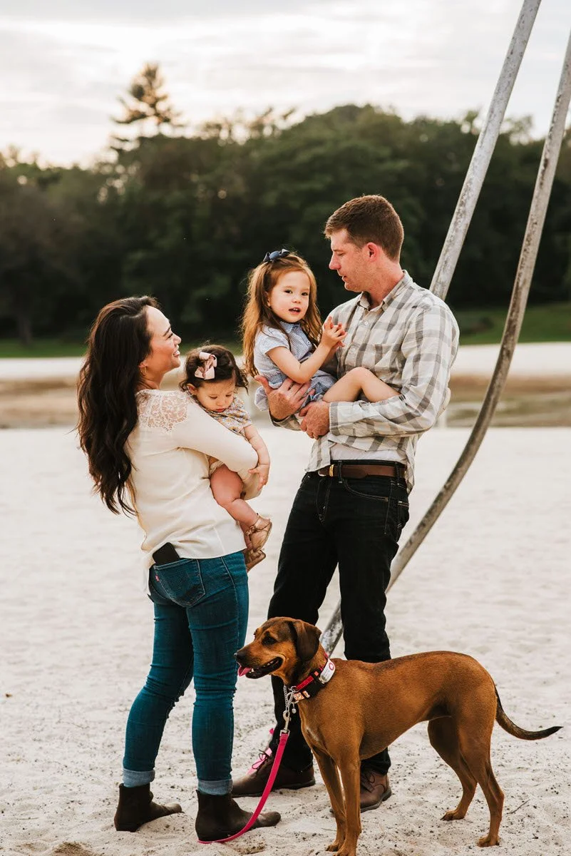 A family of four with a dog standing on a sandy outdoor area near a playground, with trees in the background. The mother holds a toddler girl, the father holds an older girl, and the dog is on a leash.