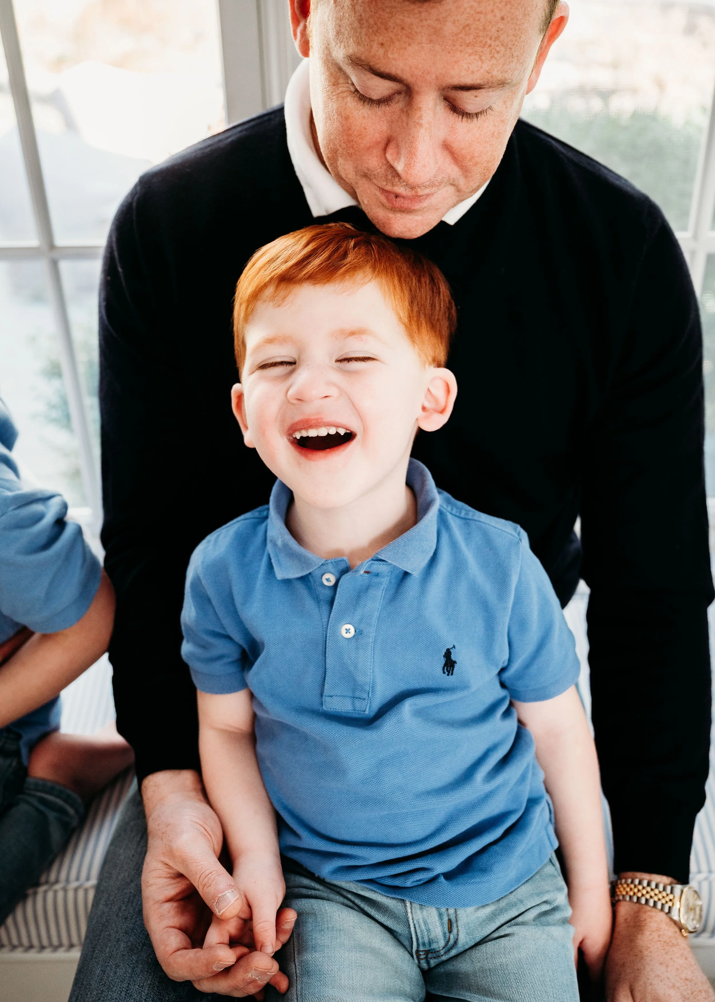 A boy with red hair smiling and laughing, sitting on someone's lap, with a man behind him leaning over and smiling.
