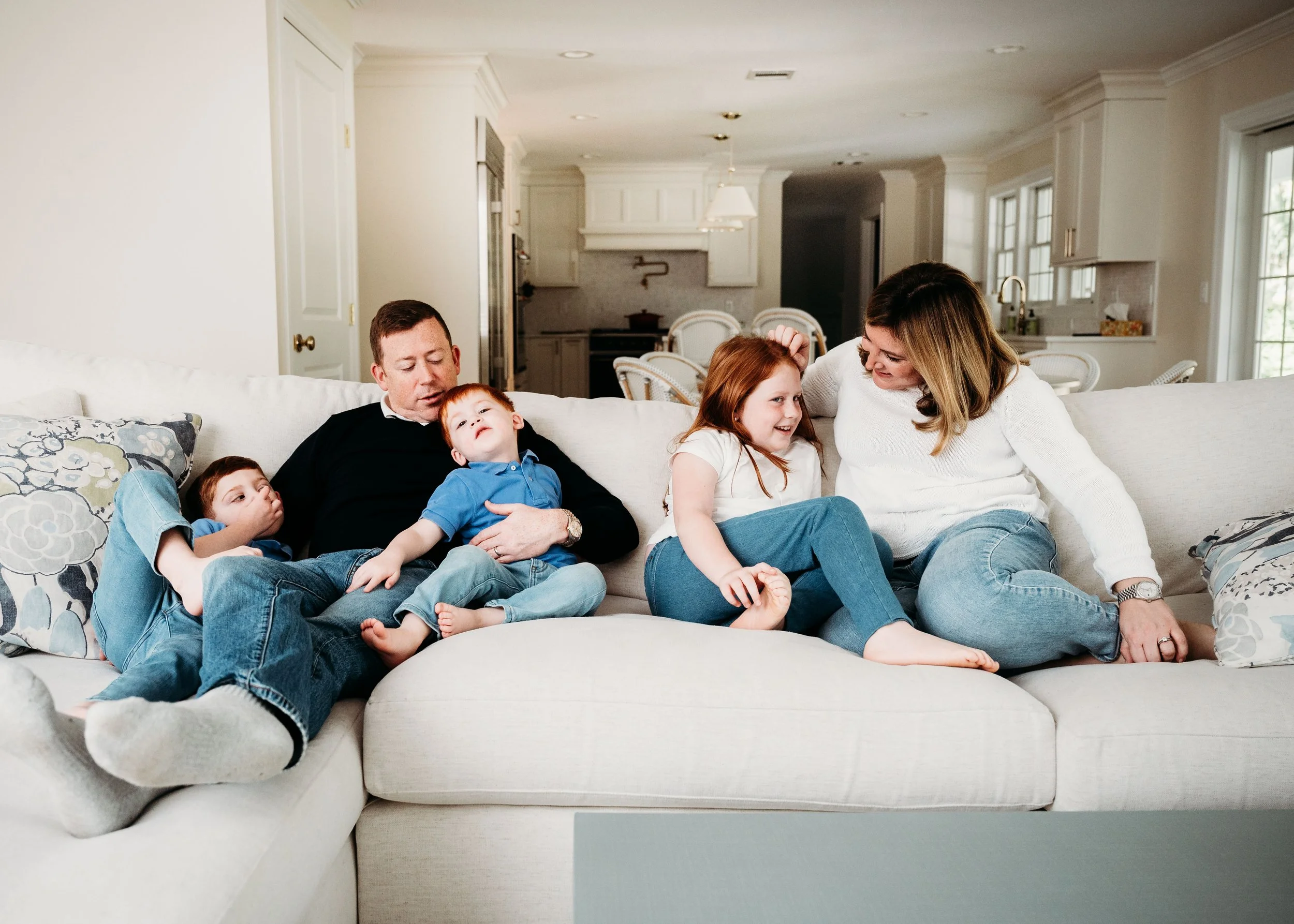 Family sitting on a white sofa in a bright living room, with children and adults, enjoying each other's company.
