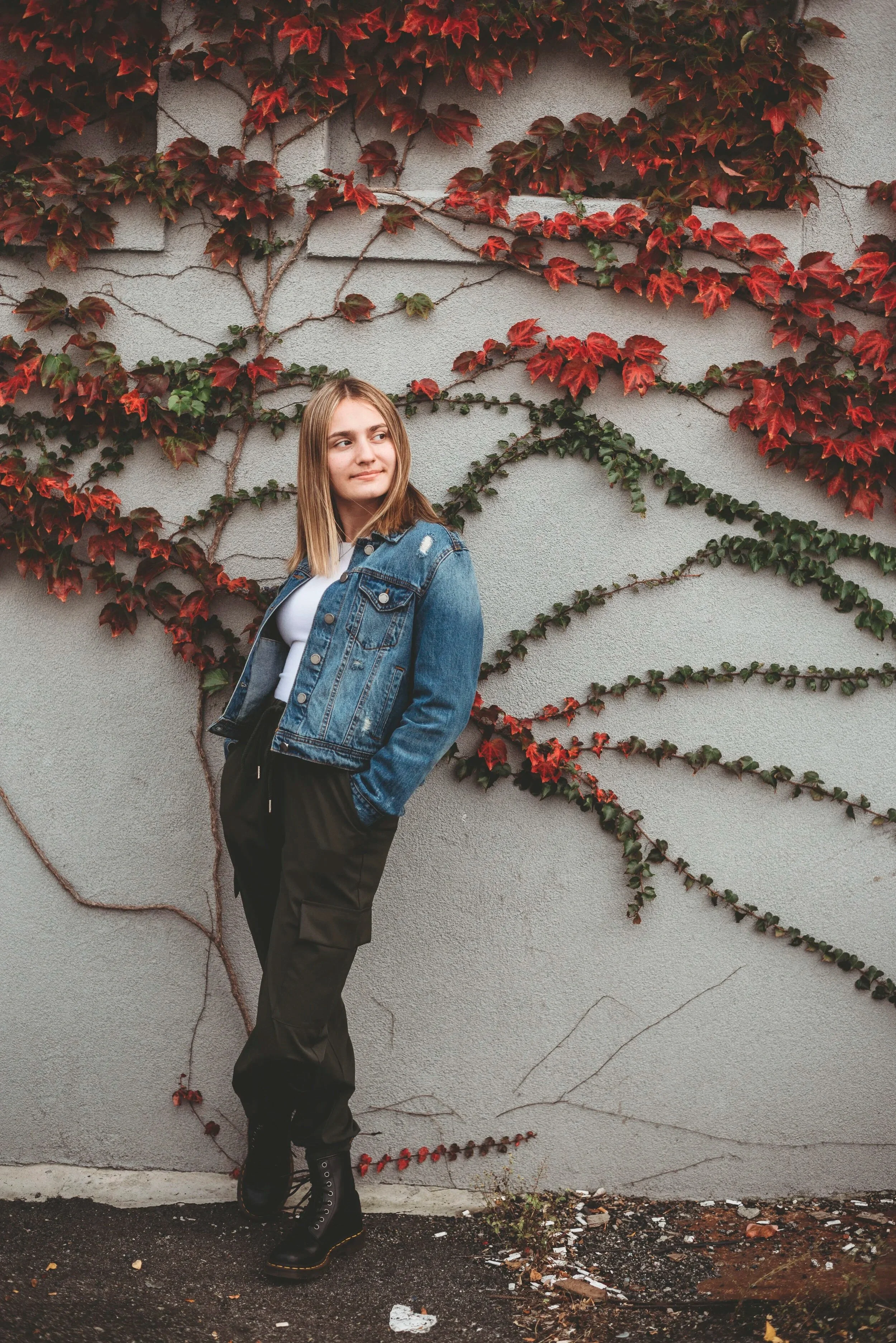 Young woman standing against a wall covered with climbing red and green ivy plants, wearing a denim jacket, white shirt, black pants, and black boots.