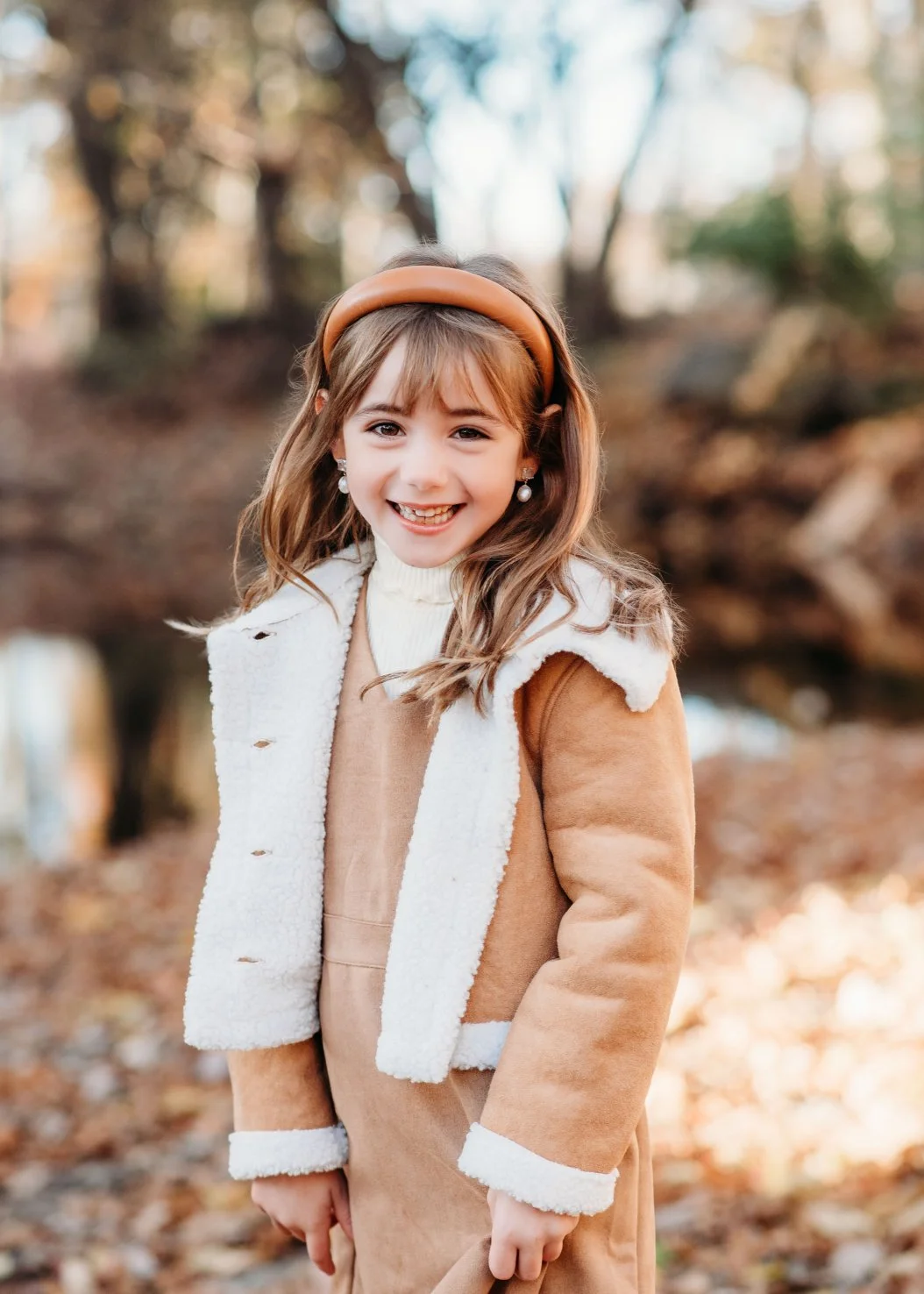 A young girl with light brown hair and a big smile, wearing a tan and white coat, a white turtleneck, pearl earrings, and a tan headband, standing outdoors in an autumnal setting with fallen leaves and blurred trees in the background.