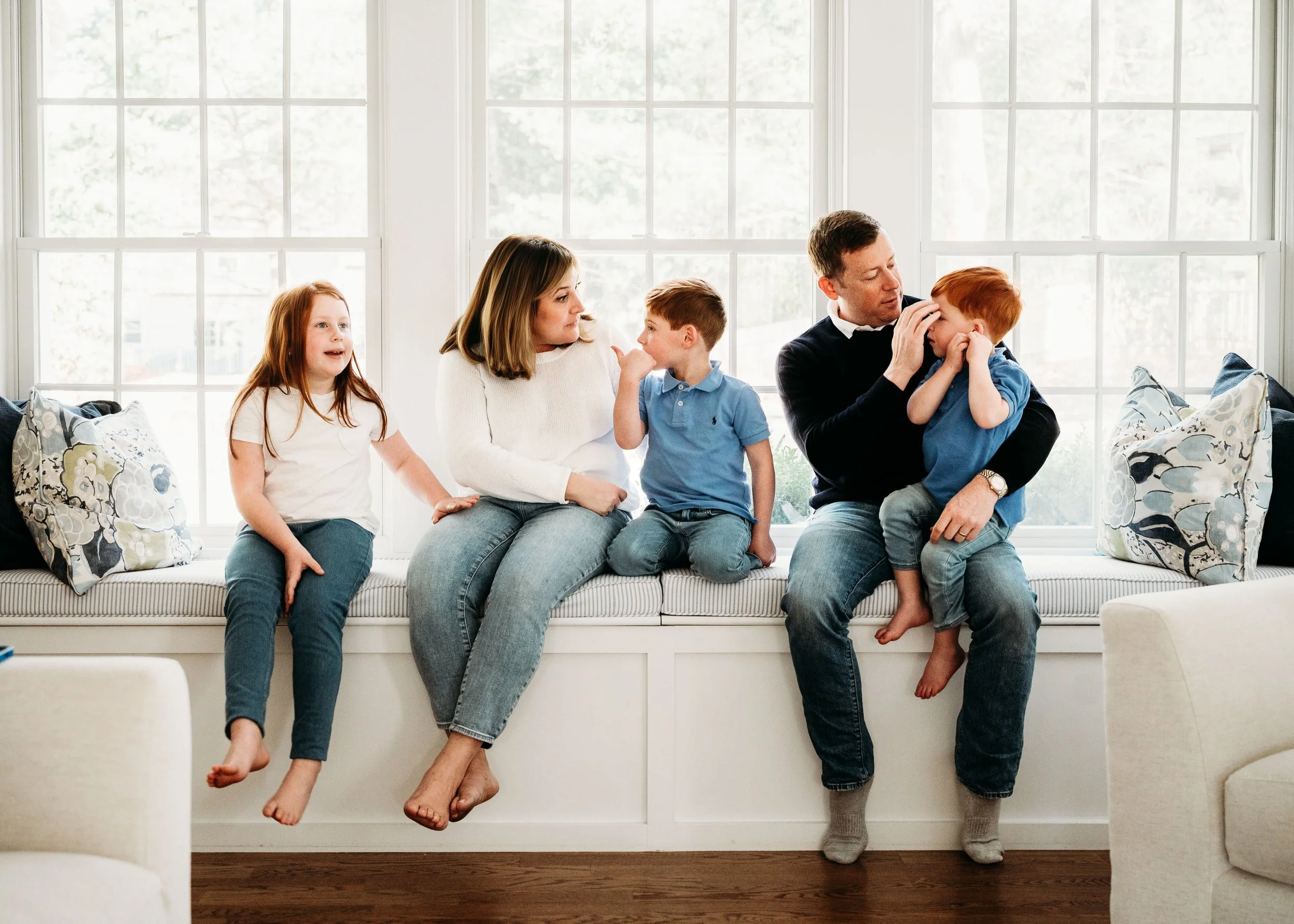 A family of five seated on a window seat in a living room, engaging in conversation and affection with each other.