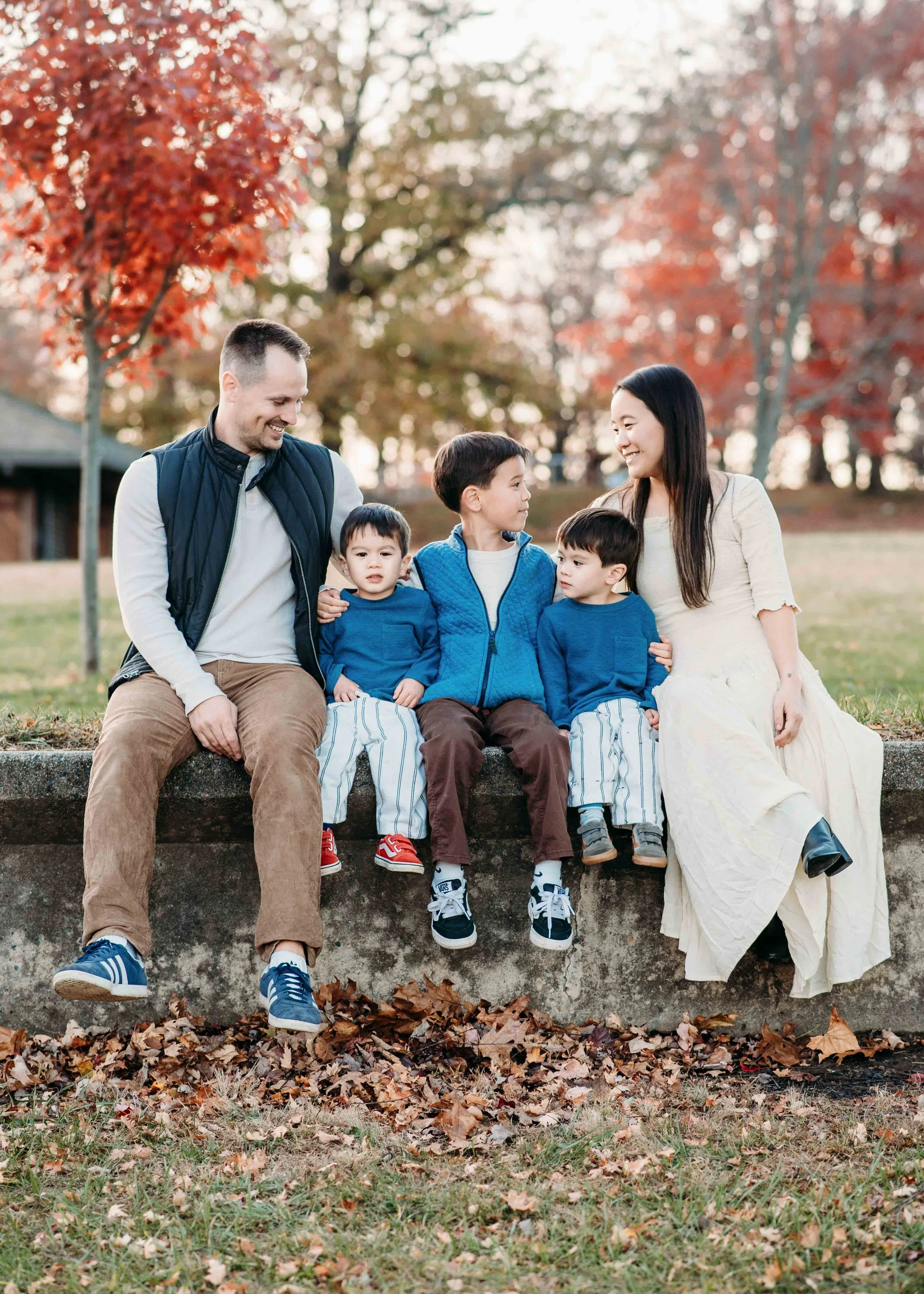A family of six sitting on a stone ledge outdoors during autumn, with colorful fall trees in the background. The adults are smiling and looking at each other, with three children sitting between them.