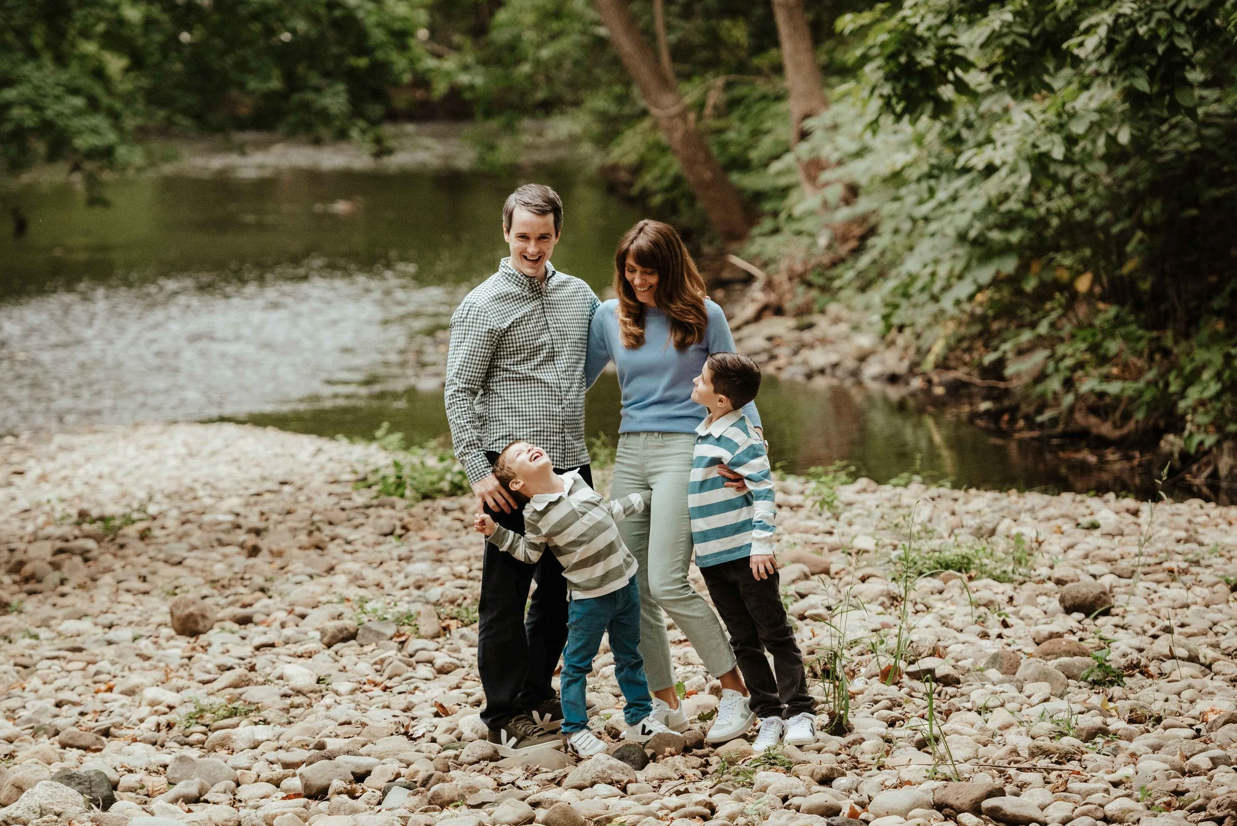 A family of four standing on a rocky riverbank, smiling and laughing, with a river and trees in the background.